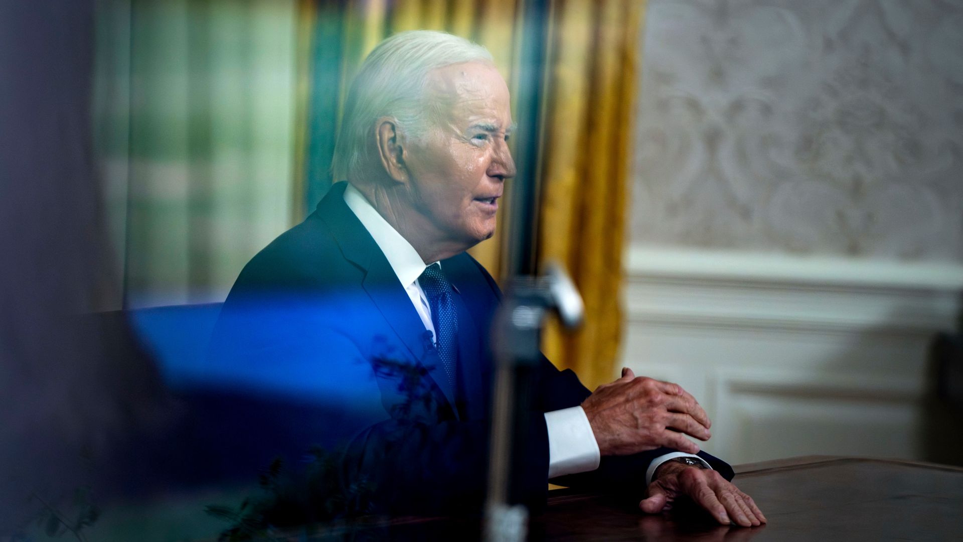 President Biden, seen through a blur, wearing a gray suit and sitting at the resolute desk in front of a gold curtain.