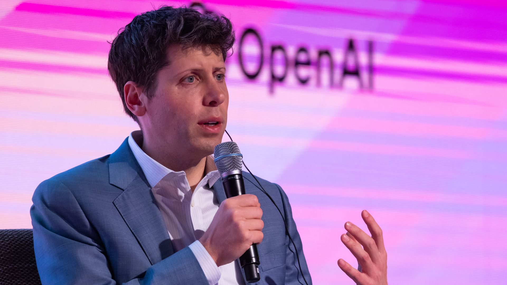 Photo of Sam Altman speaking on stage in front of sign that says "OpenAI." 
