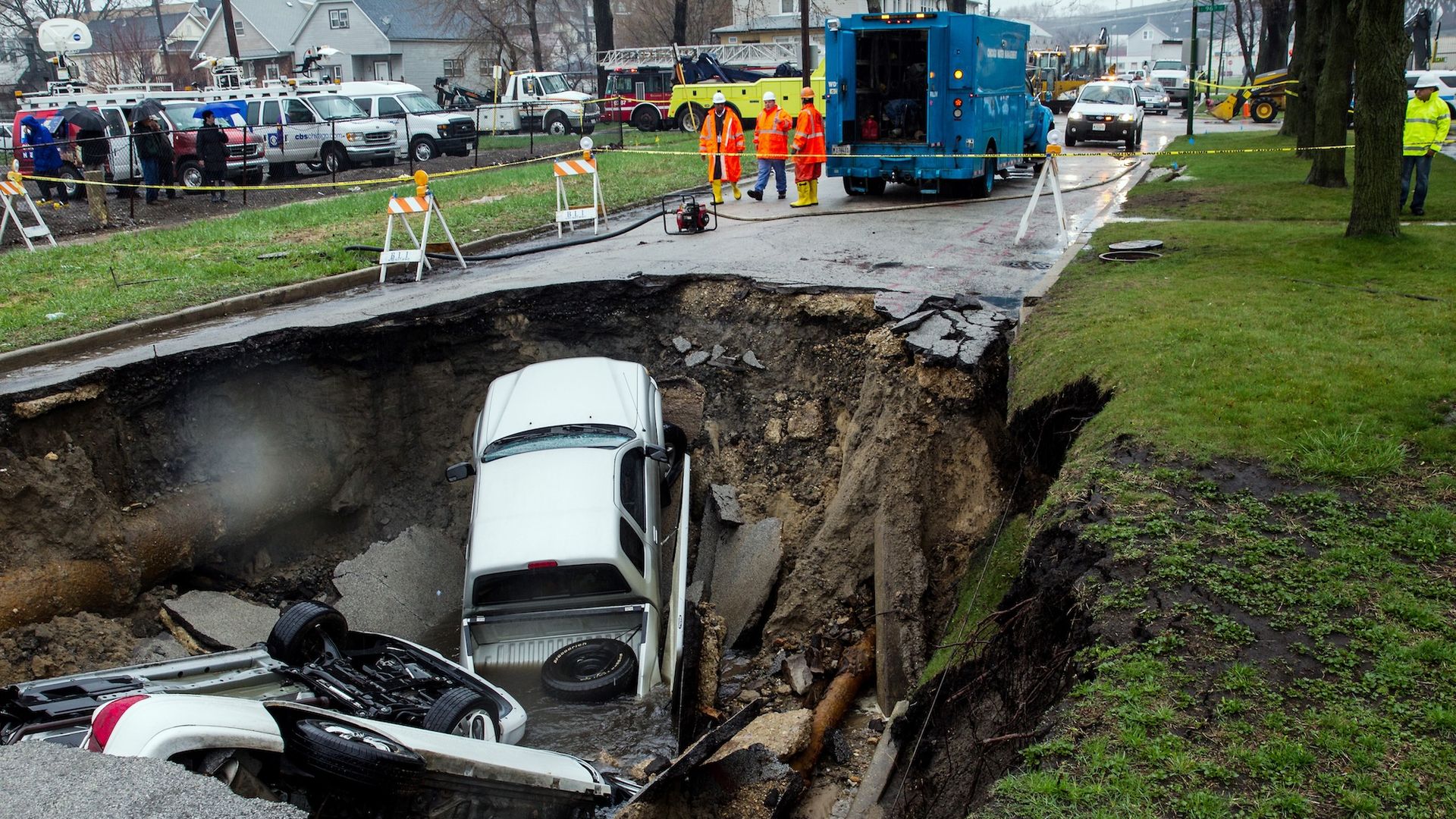 A car in a cratered sinkhole with work people surrounding it.