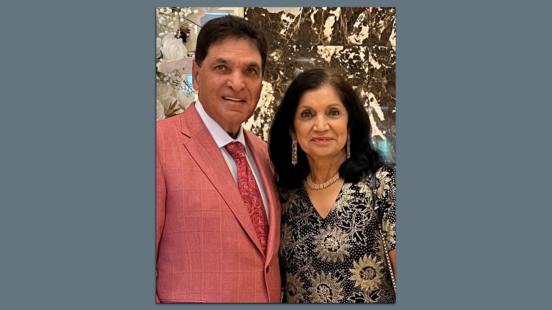 A man in a salmon-pink suit and red patterned tie stands beside a woman in a black and gold patterned dress; both smile at the camera with a marble wall backdrop.