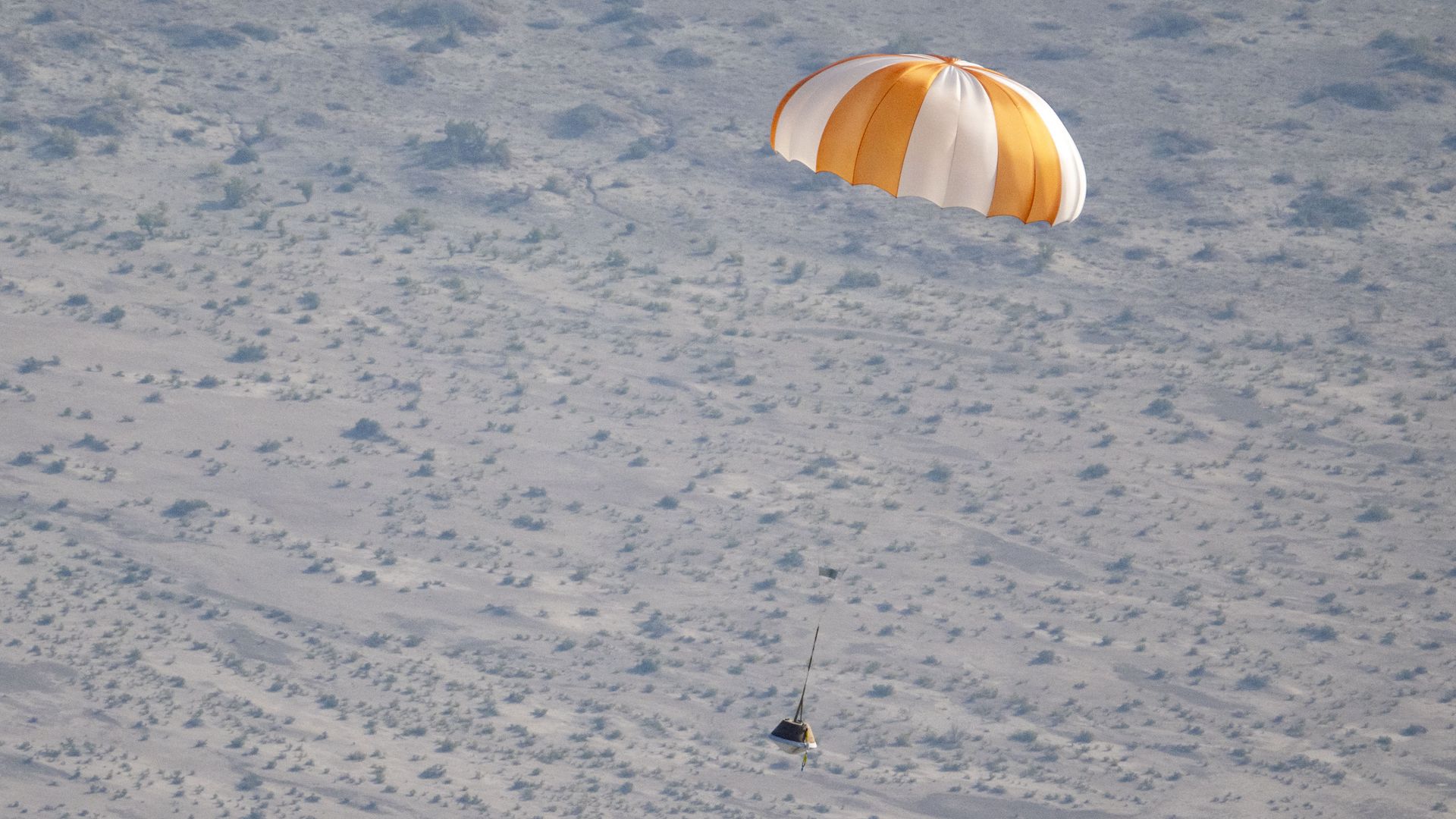 A training capsule under an orange and white parachute descends to the desert.