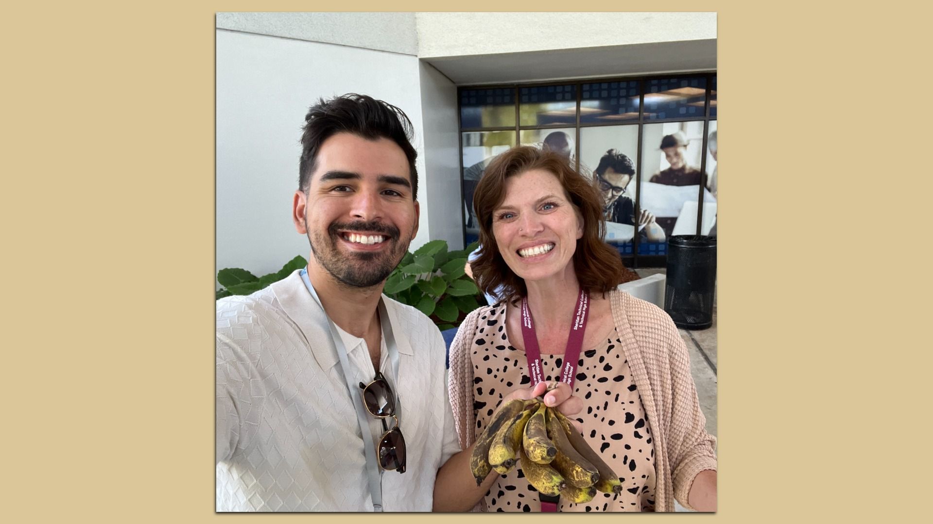 Smiling man with dark hair and a light textured shirt stands beside a smiling woman wearing a maroon lanyard; she holds a bunch of bananas. Theyโre outdoors near a building with glass windows.