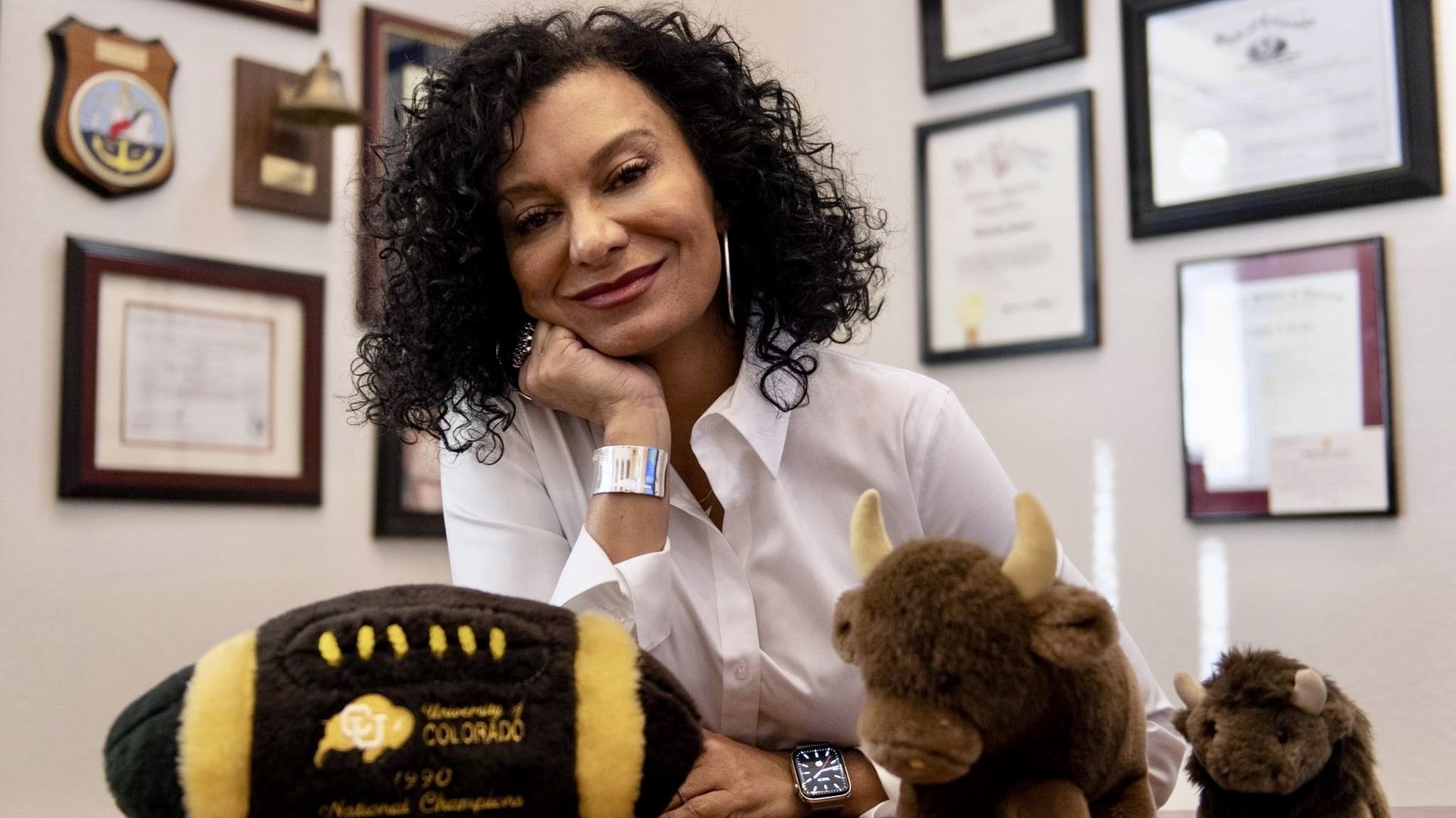 A woman with curly black hair in a white blouse sits at a desk, resting her chin on her hand. On the desk are two stuffed buffalo toys; framed certificates adorn the wall behind her.
