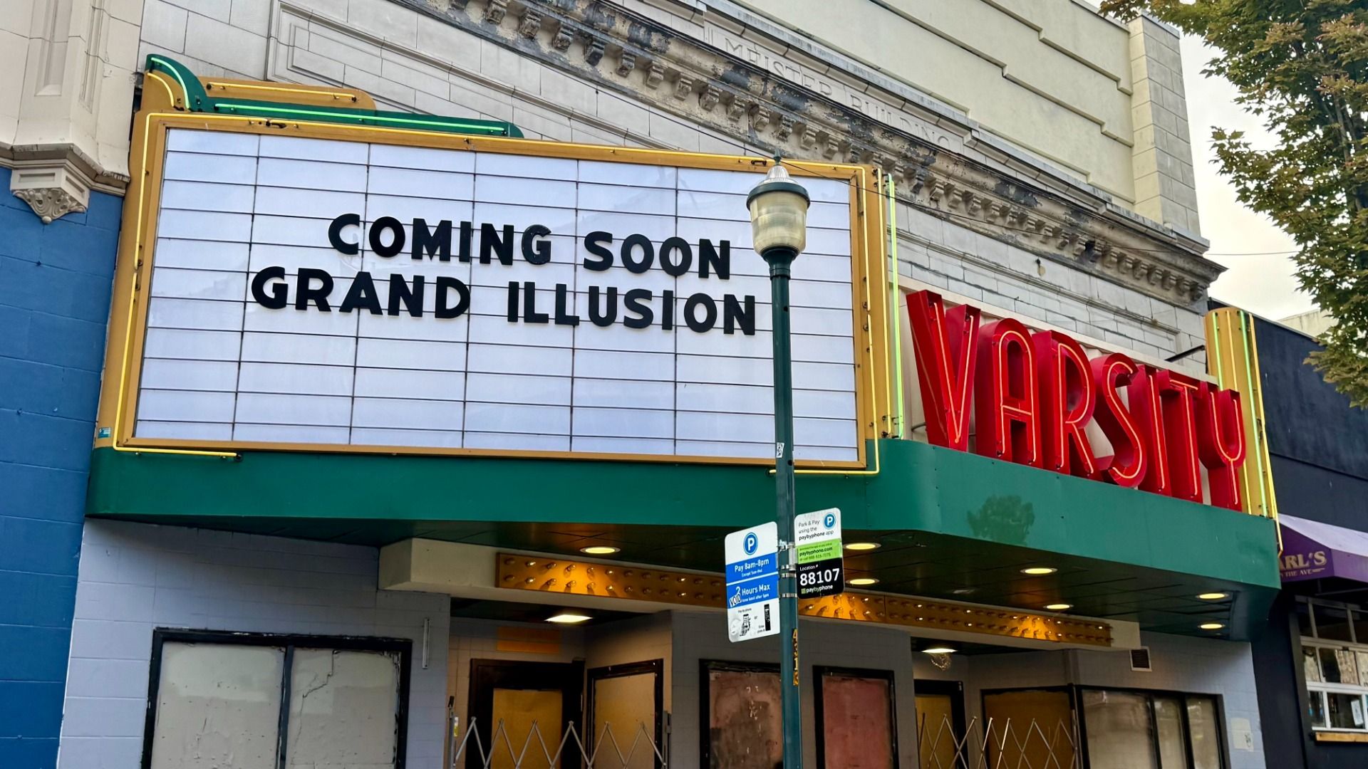 Vintage theater facade with a white marquee outlined in yellow and green reading "COMING SOON GRAND ILLUSION". Red 'VARSITY' letters on the right; a green lamppost in front; blue wall left.