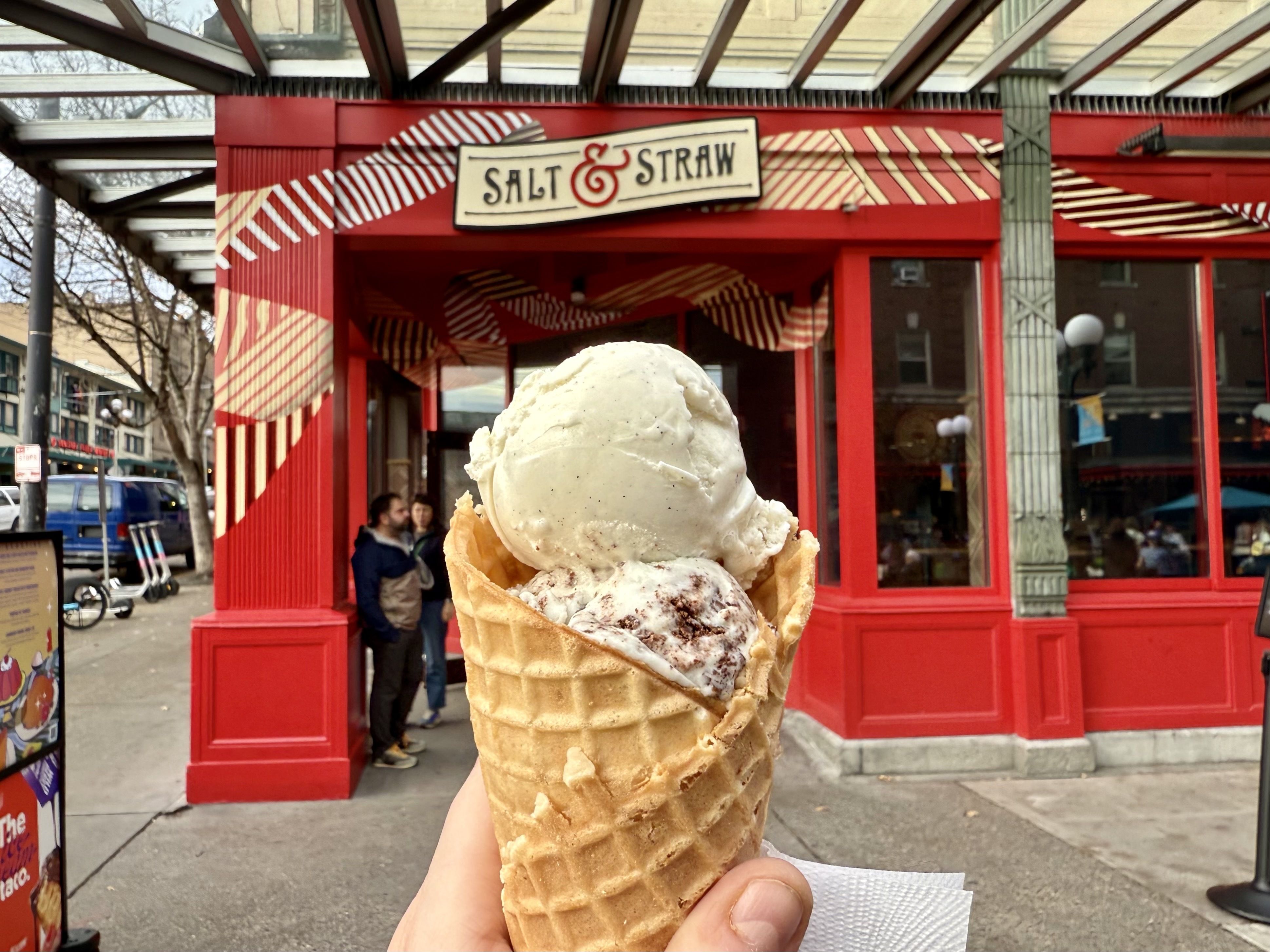 Hand holding a waffle cone with two scoops of ice cream in front of the red and beige storefront of Salt & Straw ice cream shop on a city sidewalk.
