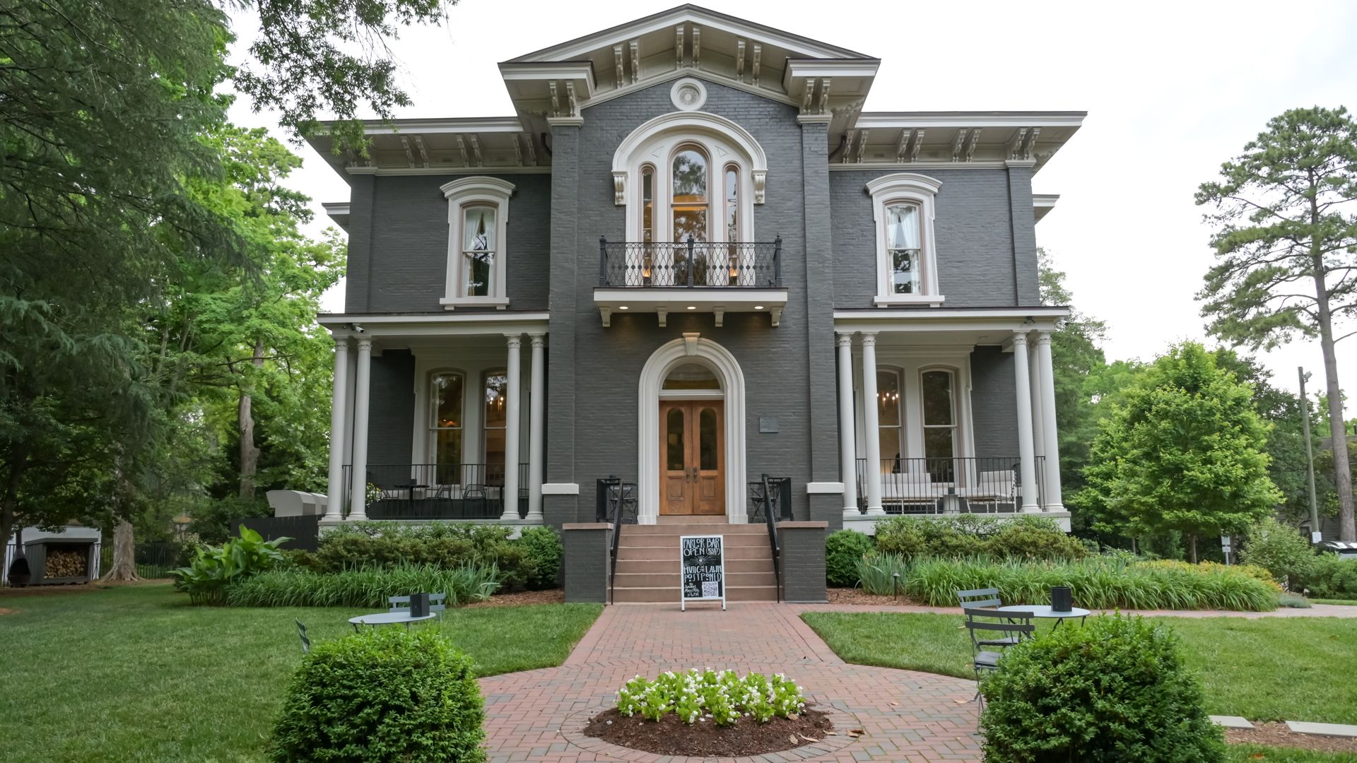 Gray brick Victorian house with white trim and a small wrought-iron balcony, flanked by tall trees. Brick path leads to wooden front doors, with columns and a chalkboard sign on the steps.