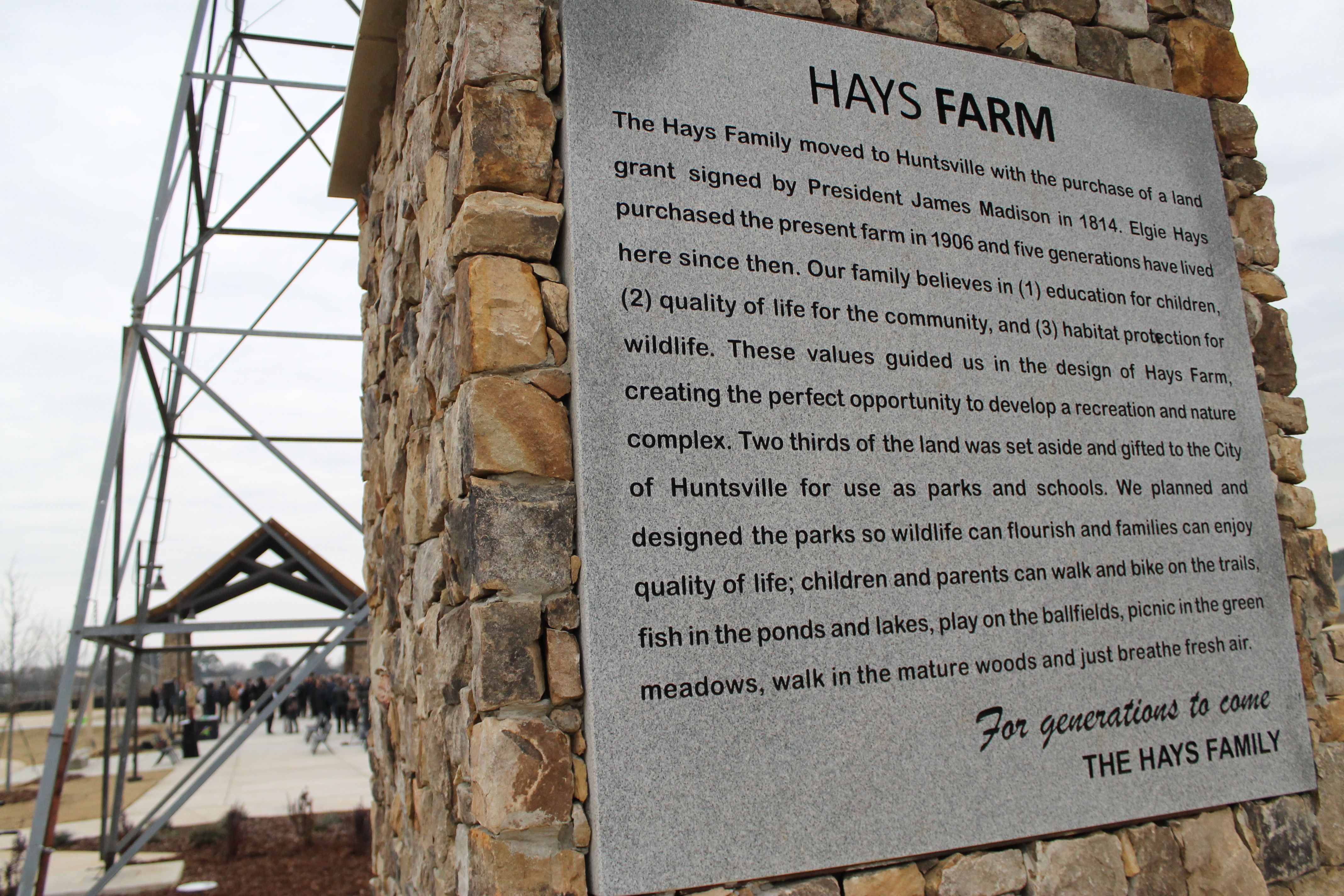 Stone monument with a plaque about Hays Farm history, family values, and land gift for parks and nature in Huntsville, with people and metal structure in the background under a cloudy sky.