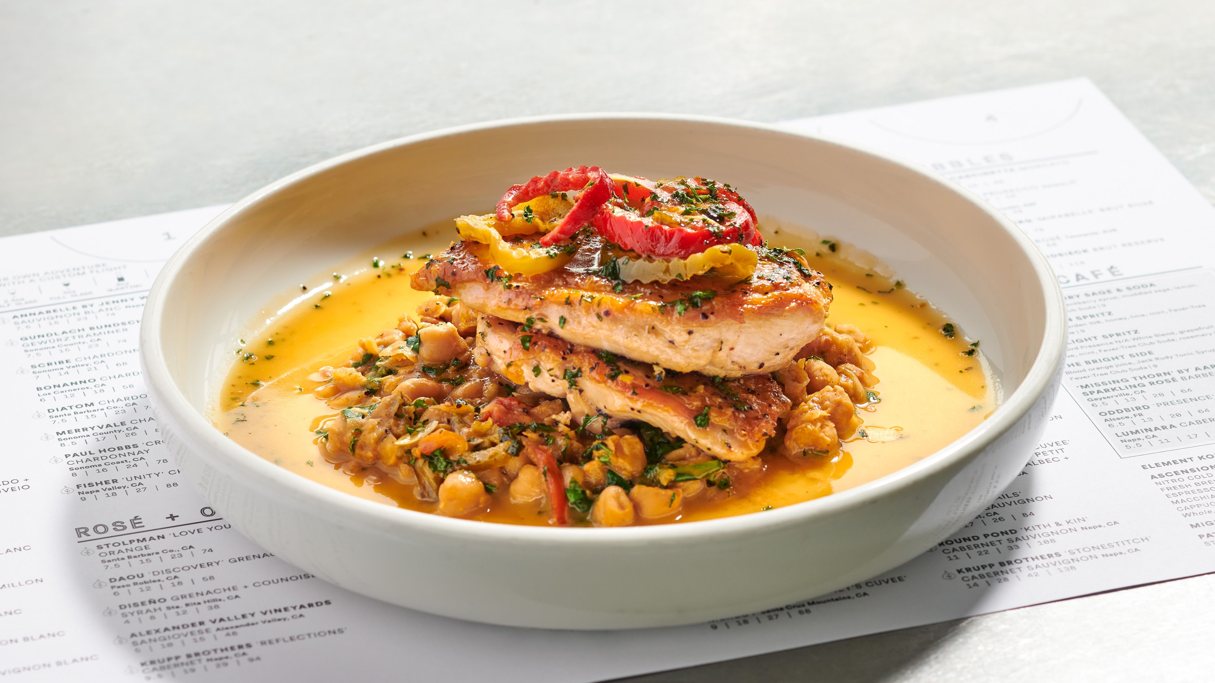 Seared chicken on a bed of chickpeas and vegetables in a golden sauce, garnished with yellow and red pepper ribbons and herbs, served in a white bowl on a menu-page backdrop.