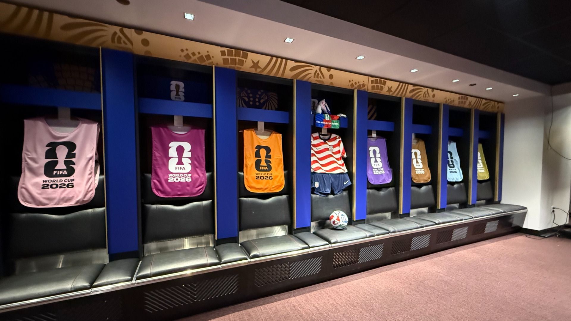 Locker room display with blue cubbies and padded benches, featuring pink, purple, orange, and striped jerseys reading "FIFA World Cup 2026" and a soccer ball resting on the bench.