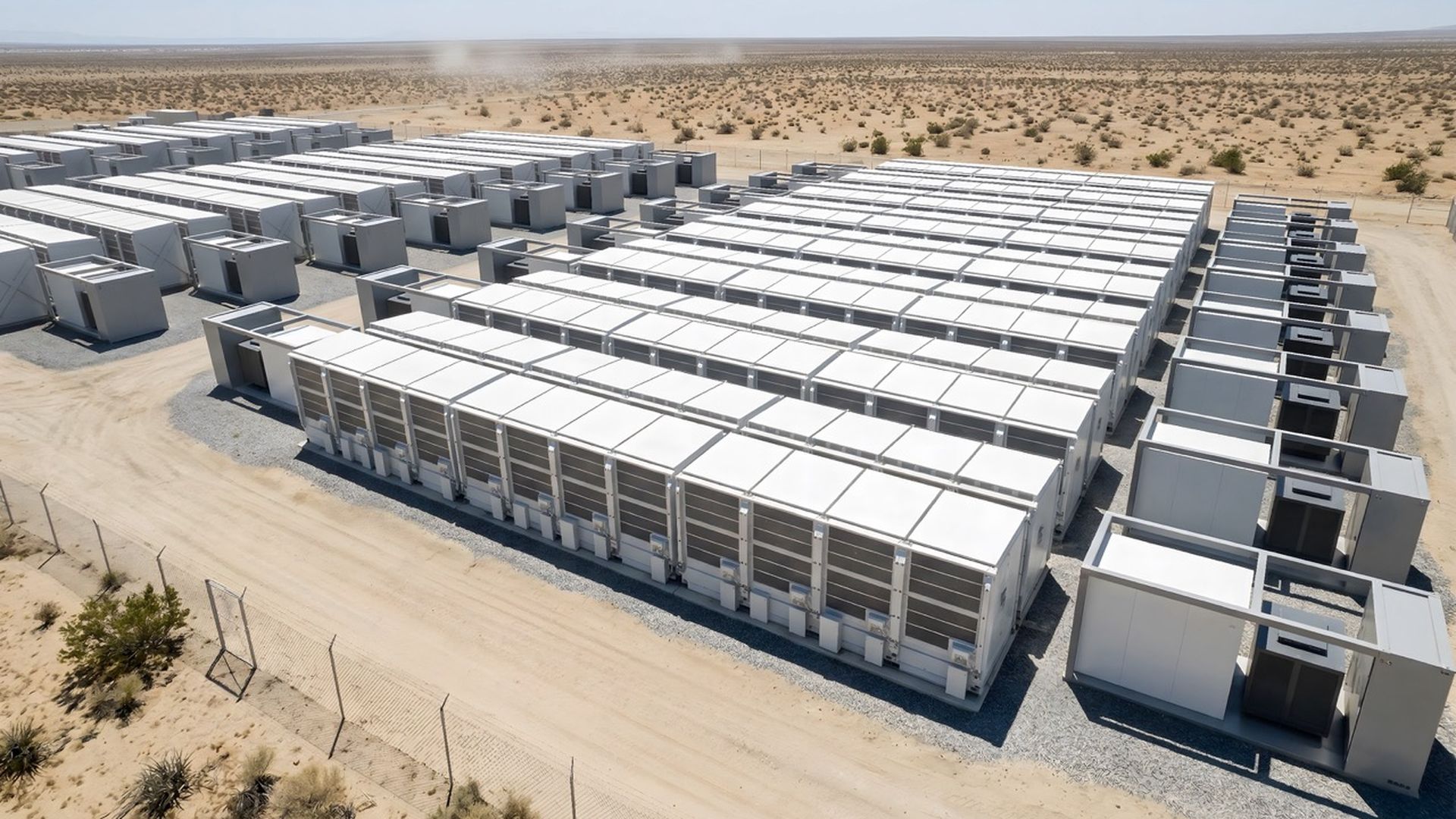 An expansive desert complex of white modular containers arranged in neat rows, likely energy storage units. Gravel roads and a perimeter fence outline the sparse landscape under a clear blue sky.