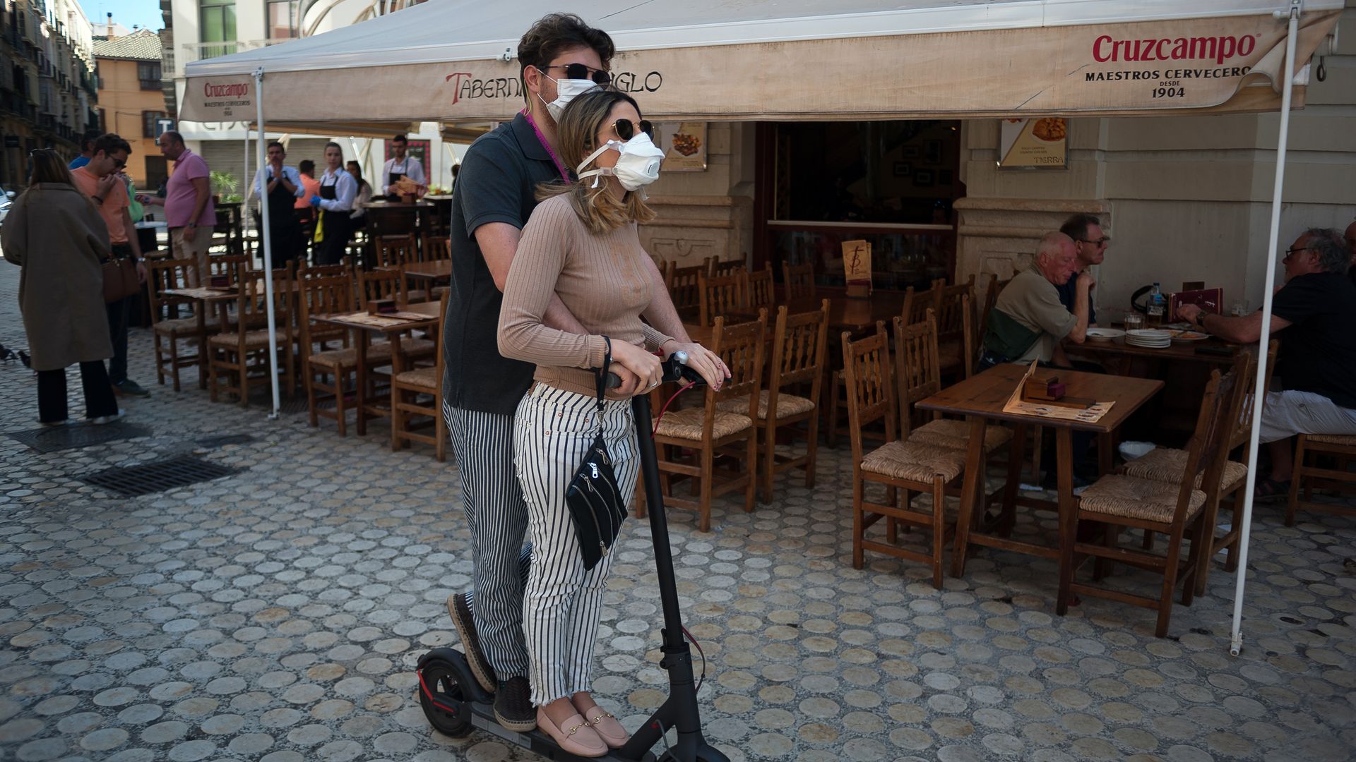 In this image, a man and a woman ride on an electric scooter while wearing face masks