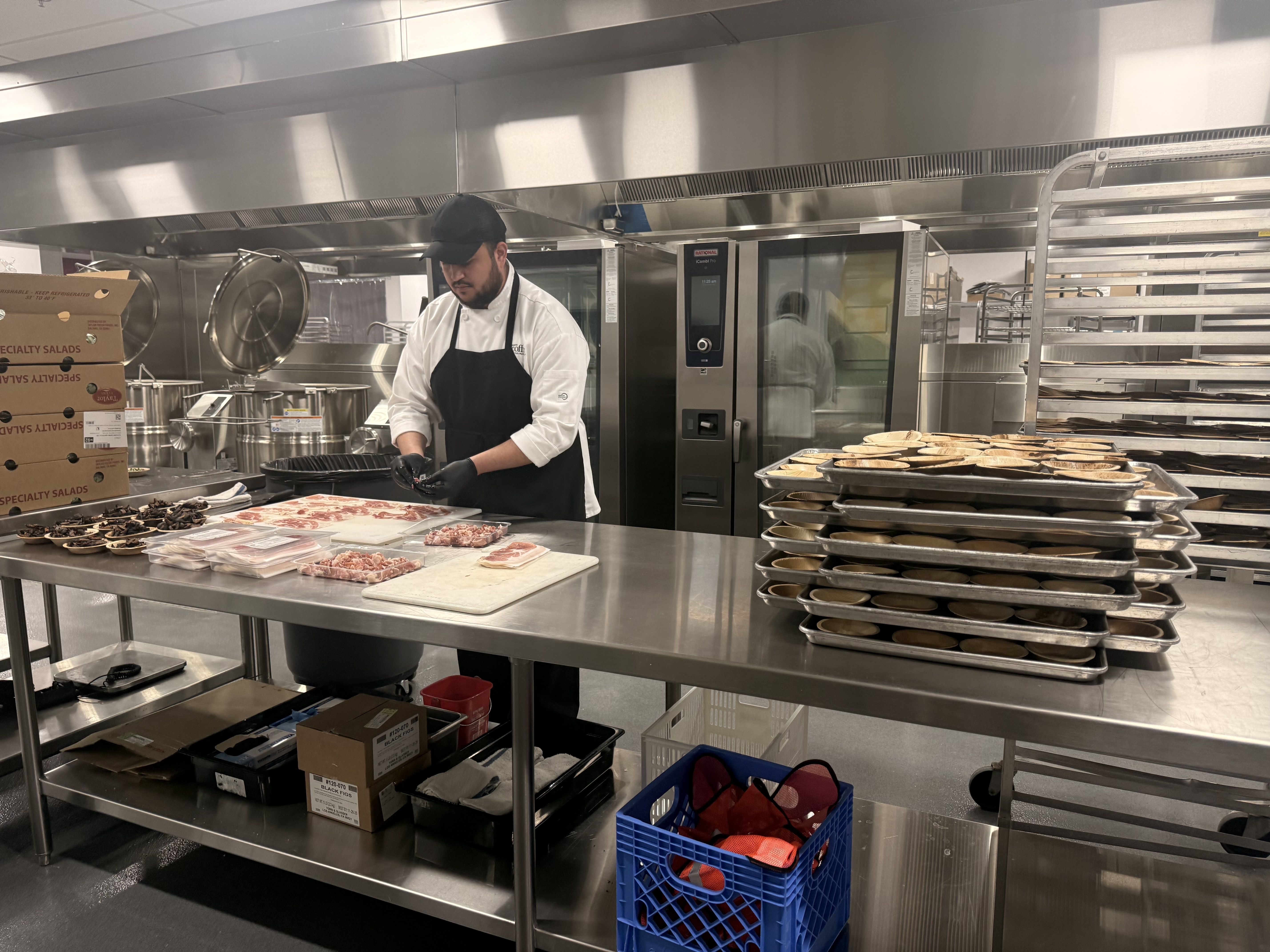 Chef in black hat and apron preparing food on a metal table in a commercial kitchen with stacked trays of baked goods and large industrial ovens in the background.