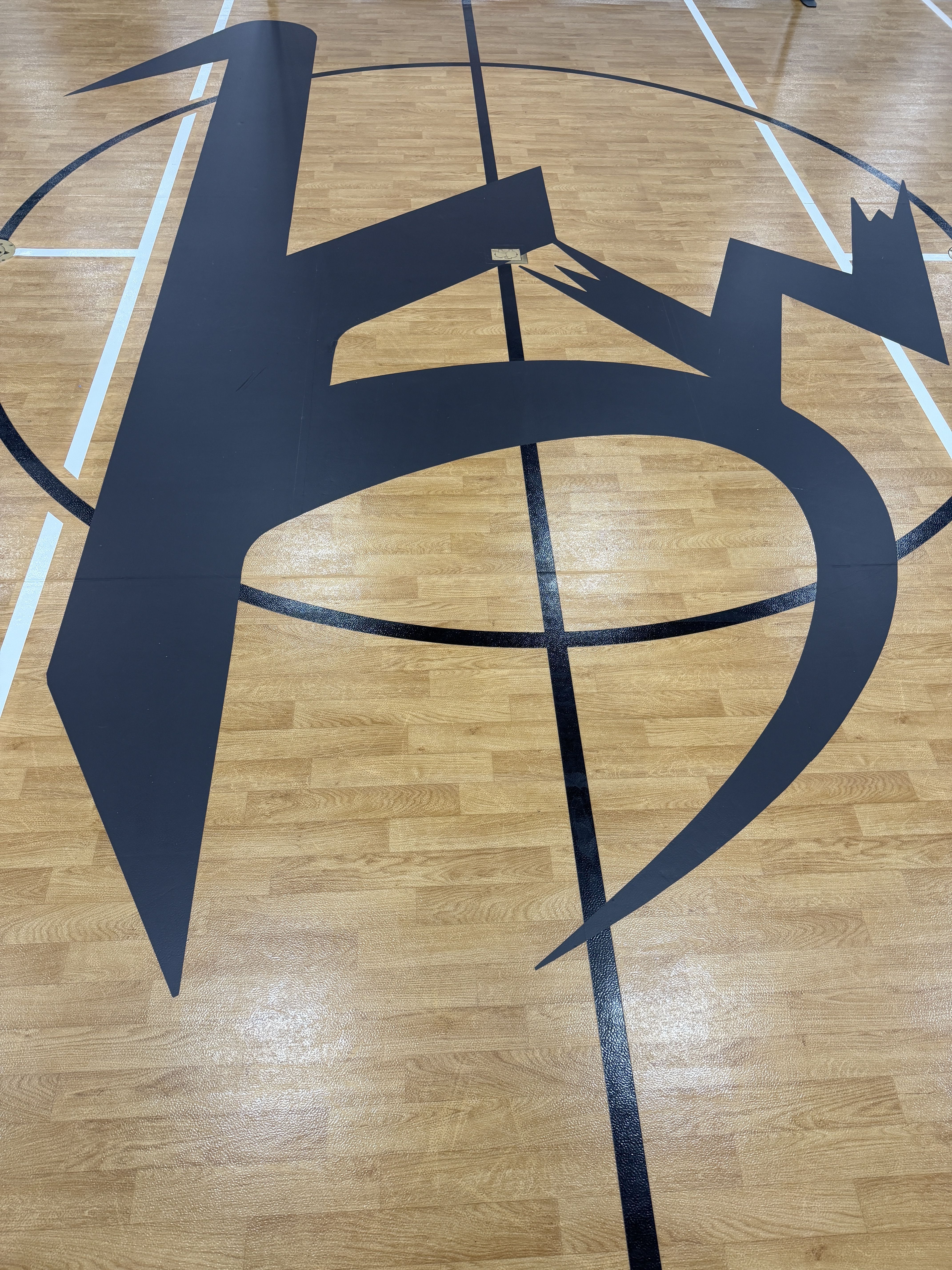 Overhead view of a gym floor with a large navy blue abstract cutout crossing the basketball court, covering lines and arcs; a small tag is attached near its center.