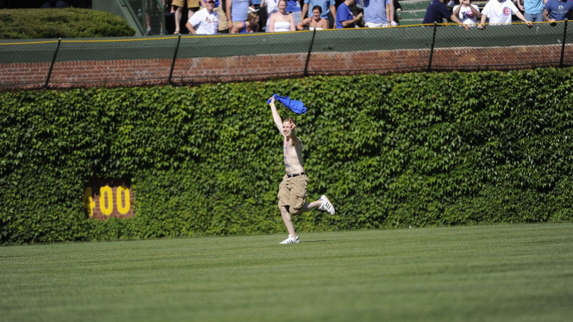 Shirtless man in tan shorts leaps on a baseball outfield, arms raised, clutching a blue shirt. A crowd in Cubs gear watches from behind a chain-link fence with ivy-covered walls.