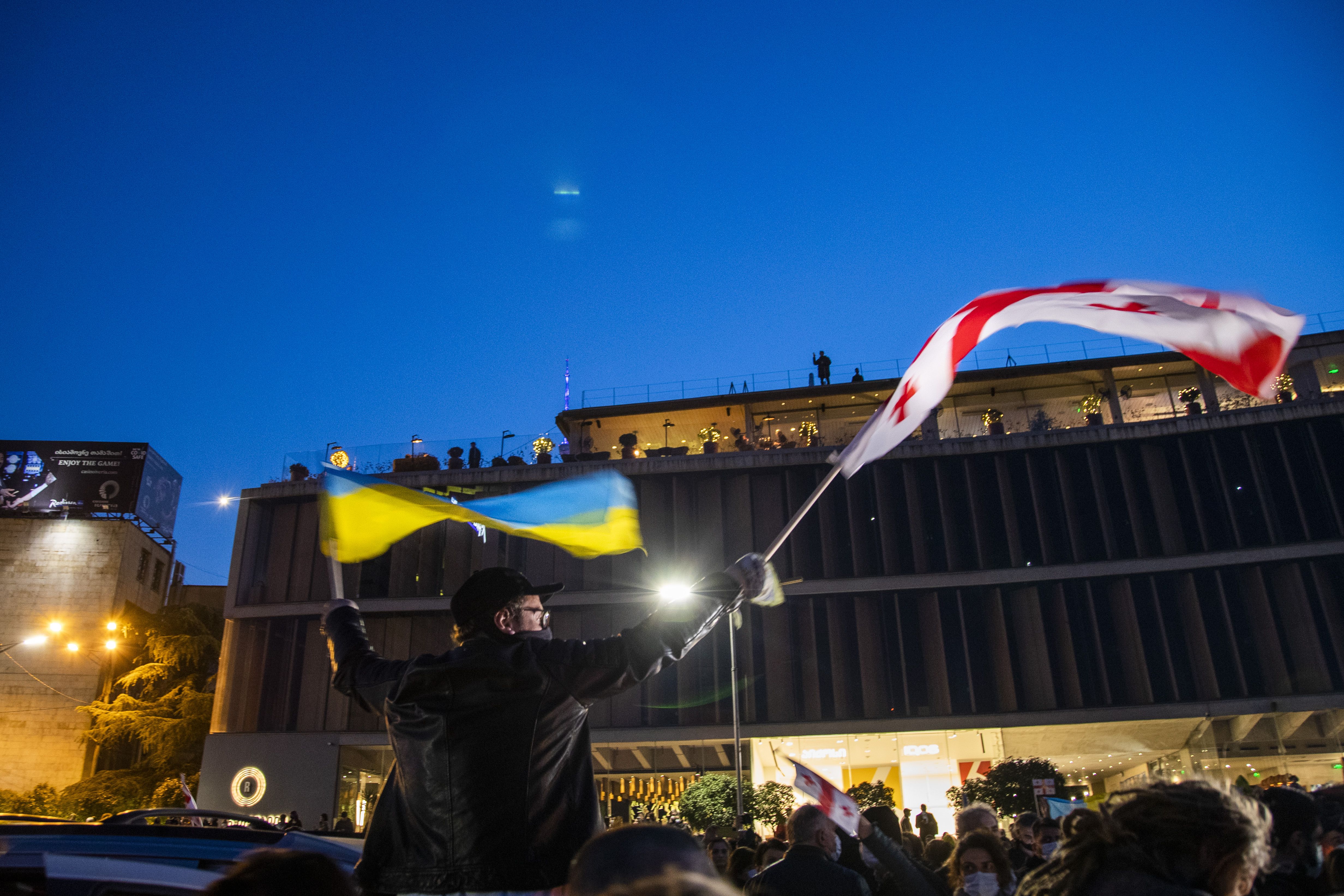 Photo of a person in a truck waving a Ukraine flag in their left hand and a Georgian flag in their right at a protest at night