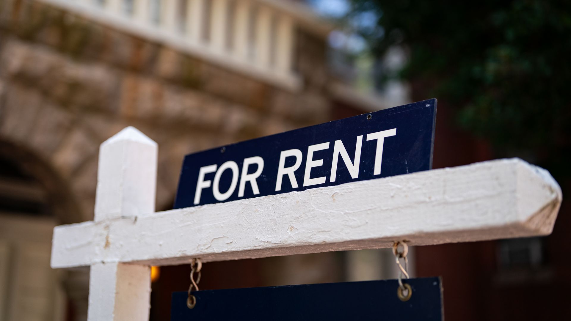 A white wooden frame in the foreground supports a dark blue sign reading FOR RENT. The background shows a stone wall, railing, and greenery with a house blurred in the distance.