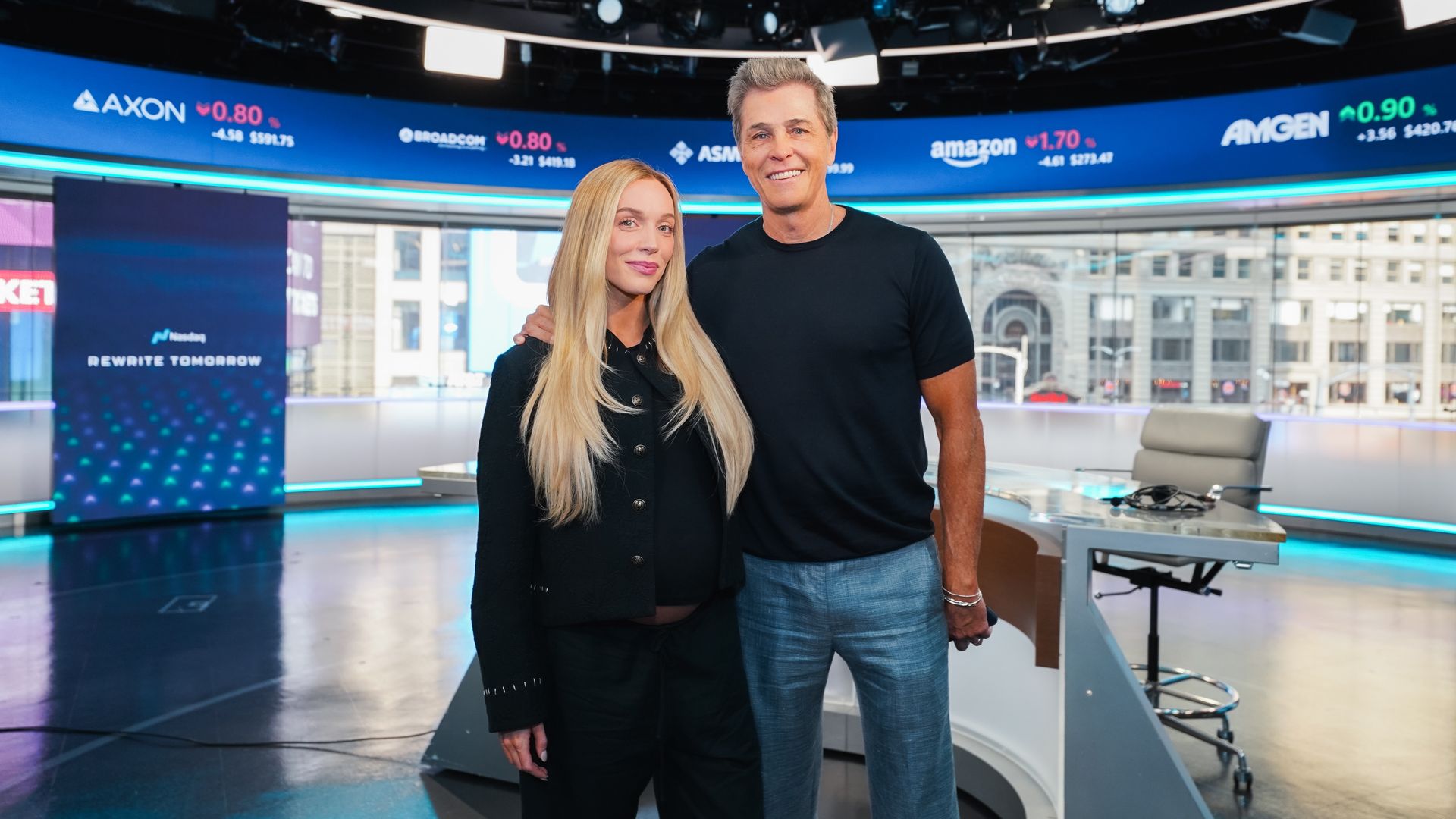 Two hosts stand in a TV studio, smiling. A curved blue ticker wall behind them shows stock quotes and logos, as bright studio lights glow and the city is visible through the windows.