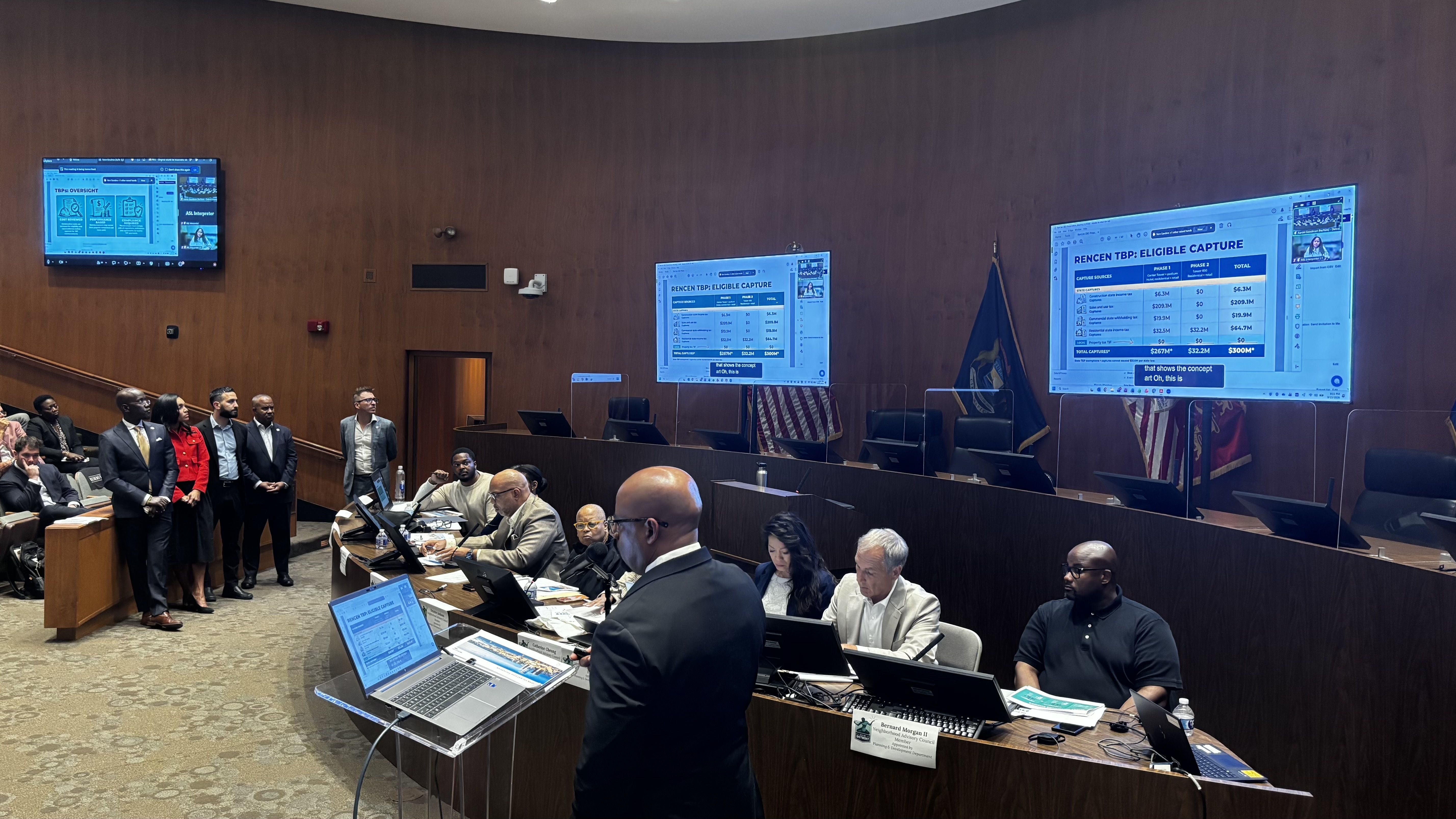 Wood-paneled conference room with large screens showing a spreadsheet titled "RENCEN TBP: ELIGIBLE CAPTURE". A group of suited people stands left; others work at desks with laptops; flags visible.