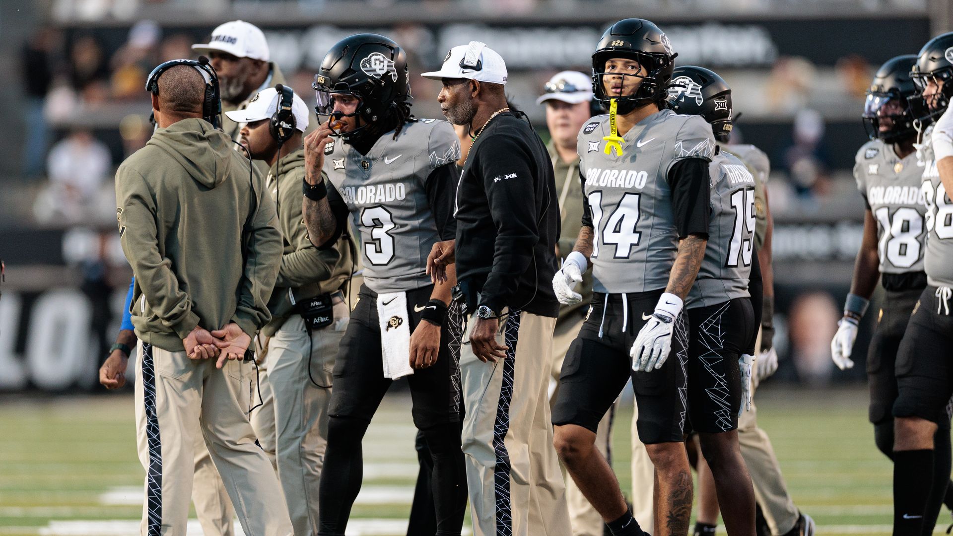 Football players in gray and black Colorado uniforms with helmets stand on a field with Deion Sanders during a game.
