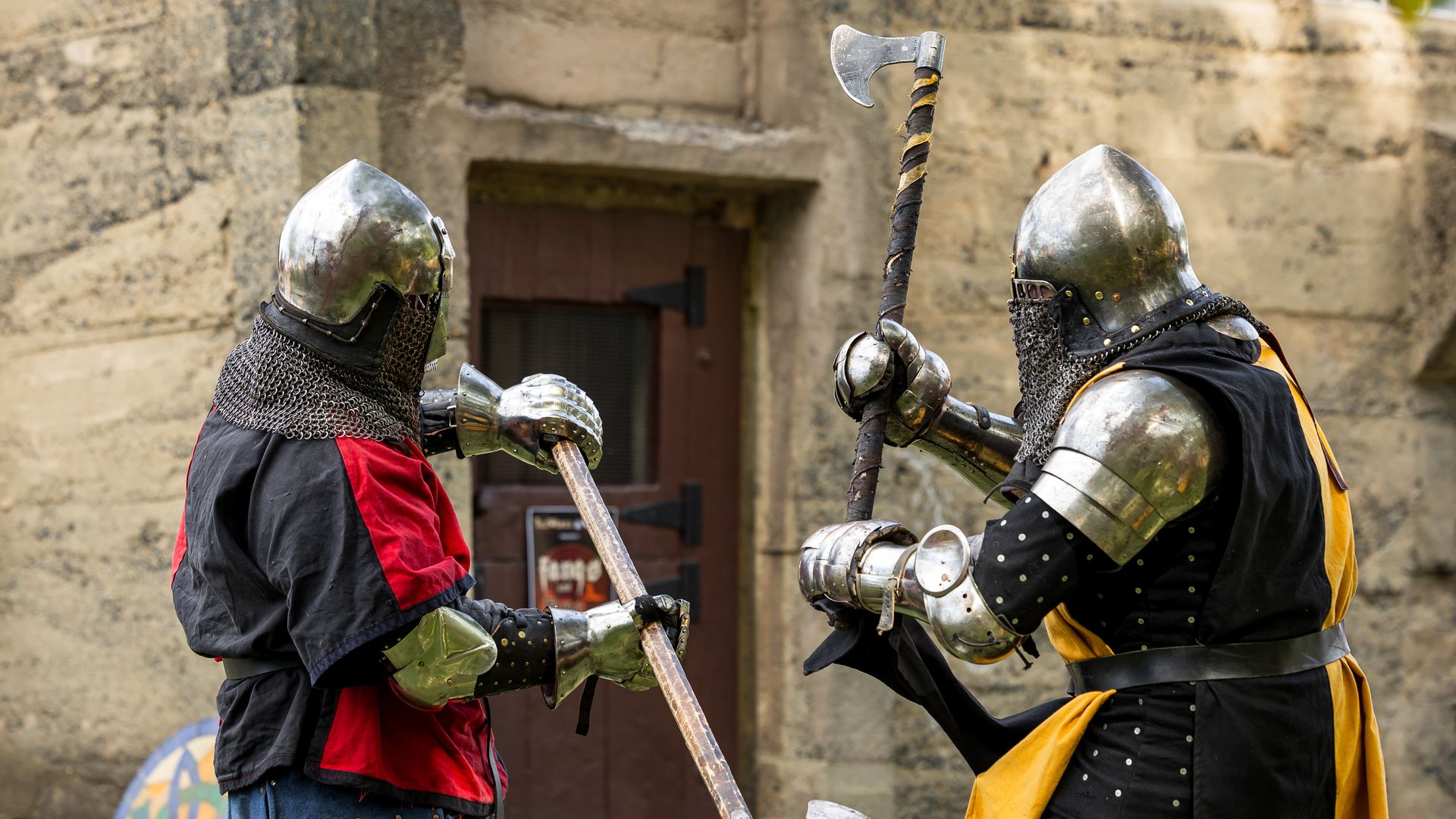 Two armored knights duel with axes in a stone courtyard; one wears red and black, the other black and yellow. Chainmail, polished helmets, and a painted round shield are visible.