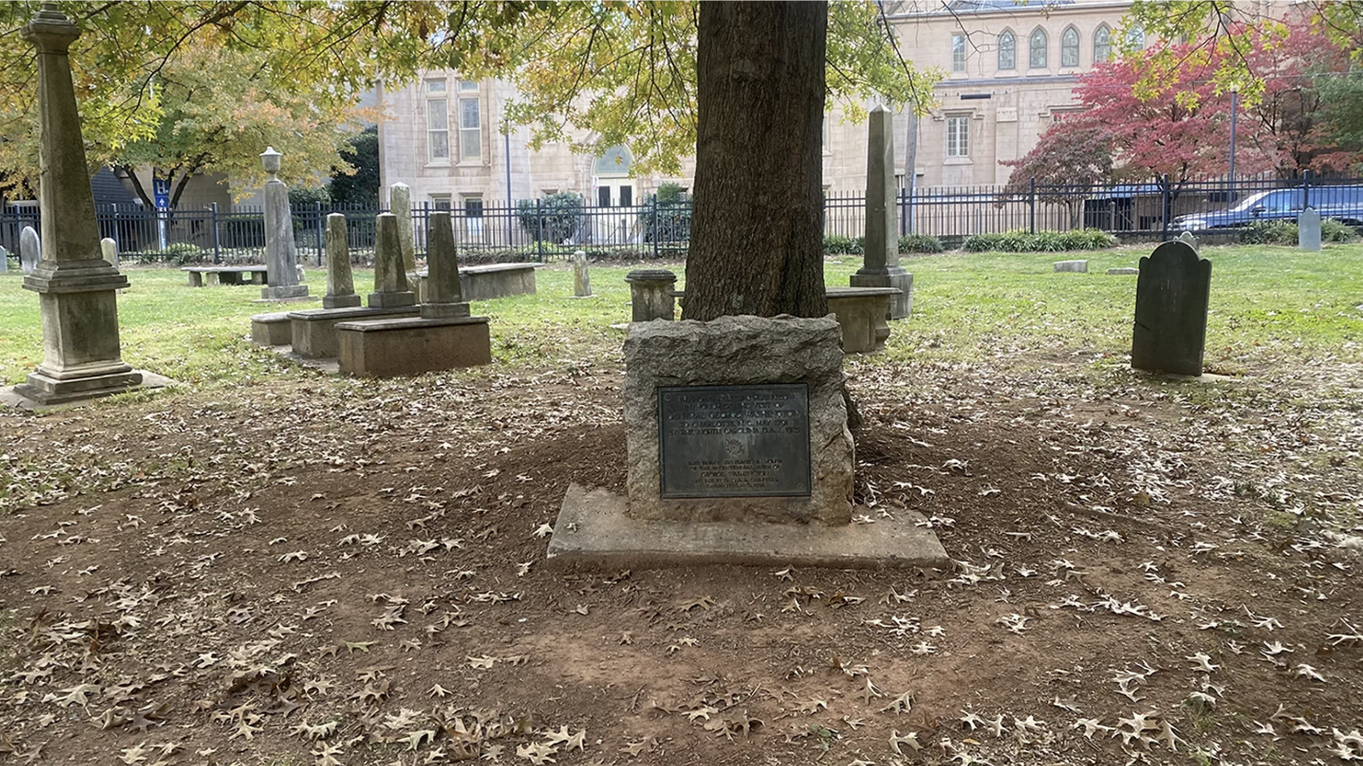A historic cemetery with stone tombstones and monuments around a large tree with a plaque, autumn leaves on the ground, green grass, and a beige building with arched windows in the background.