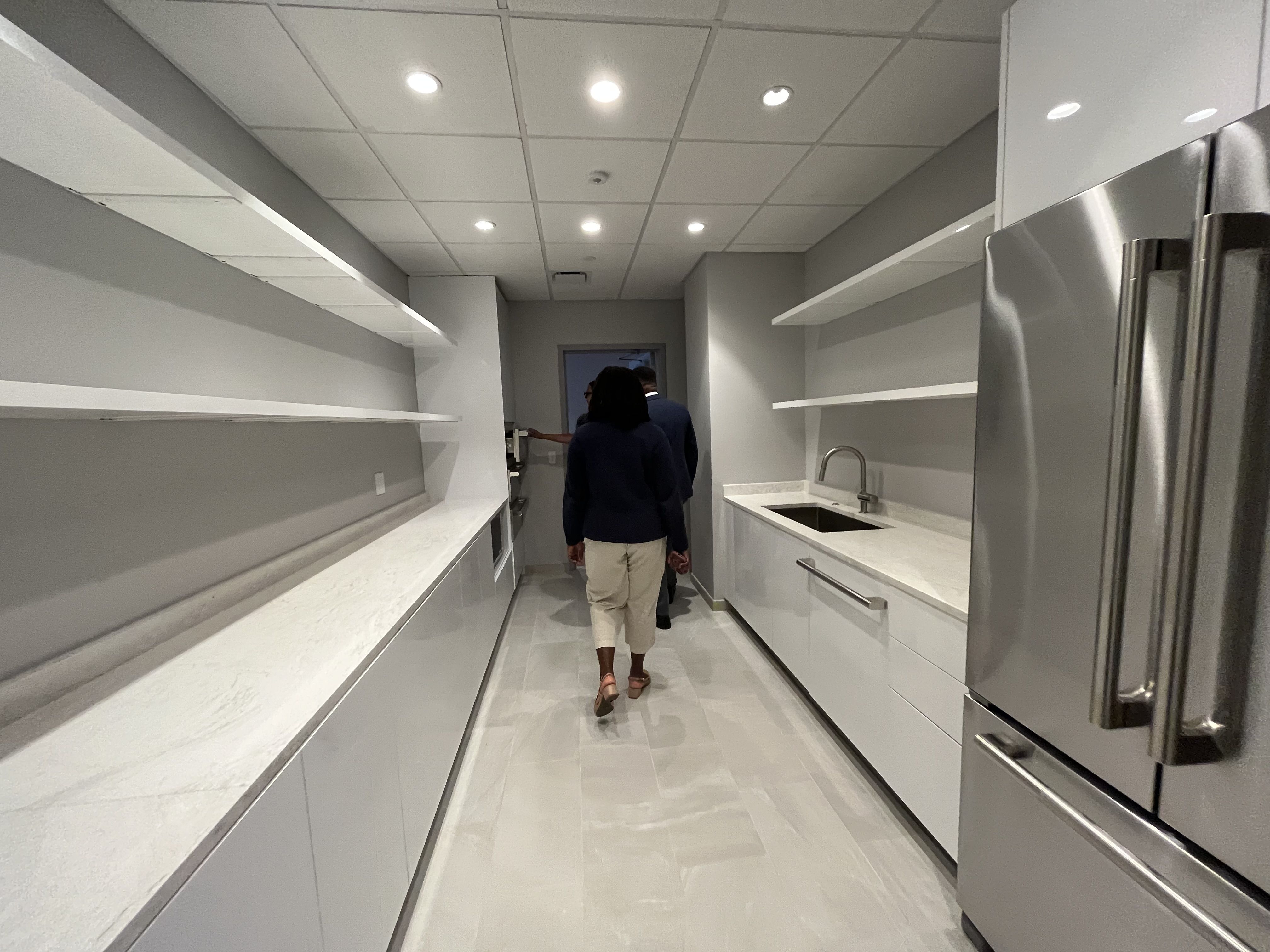 Modern narrow kitchen with white countertops, gray walls, open shelves, stainless steel refrigerator, sink on right side, and two people walking toward a doorway at the end.