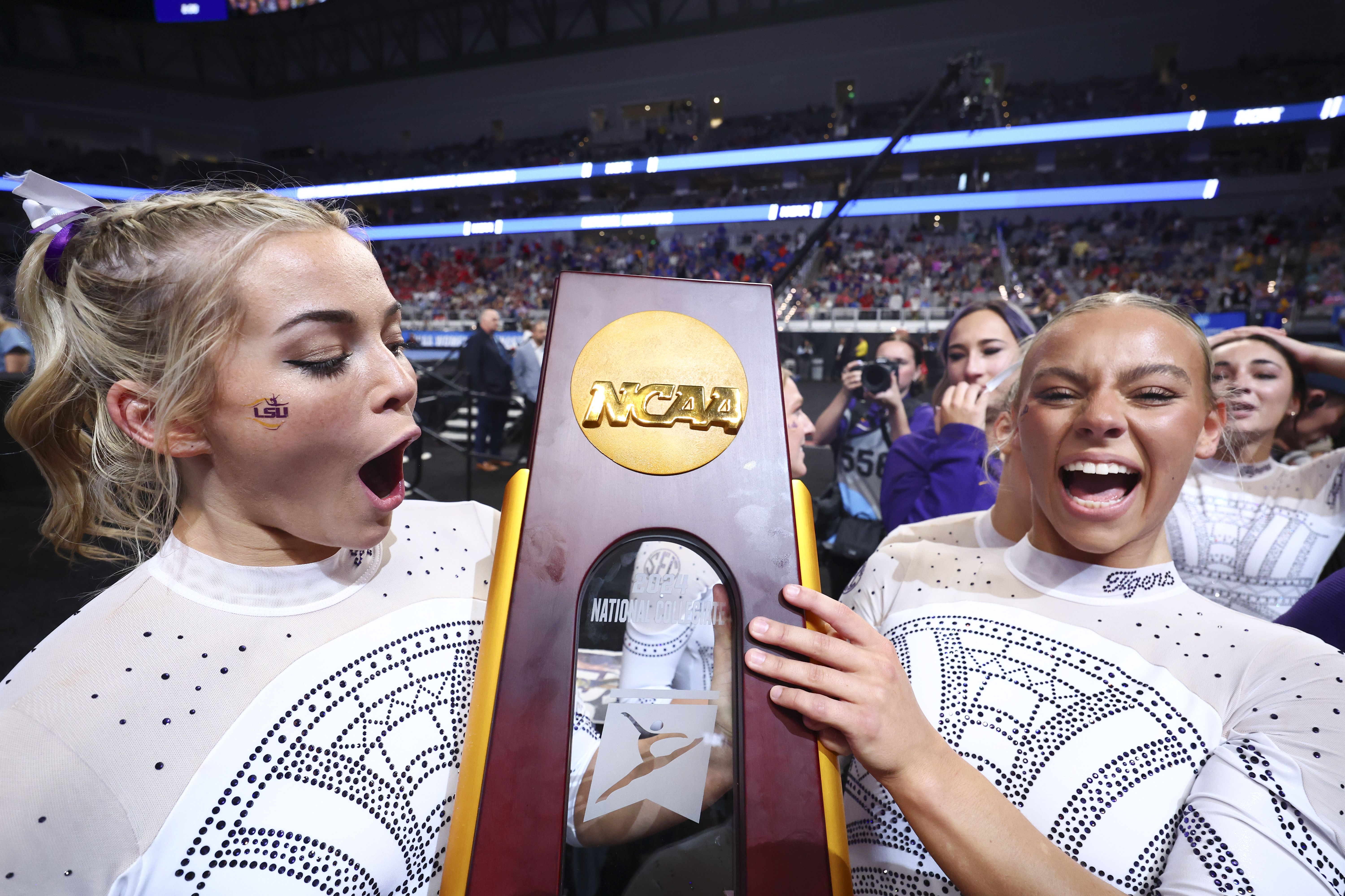 Two gymnasts hold up the NCAA gymnastics national championship trophy in celebration.