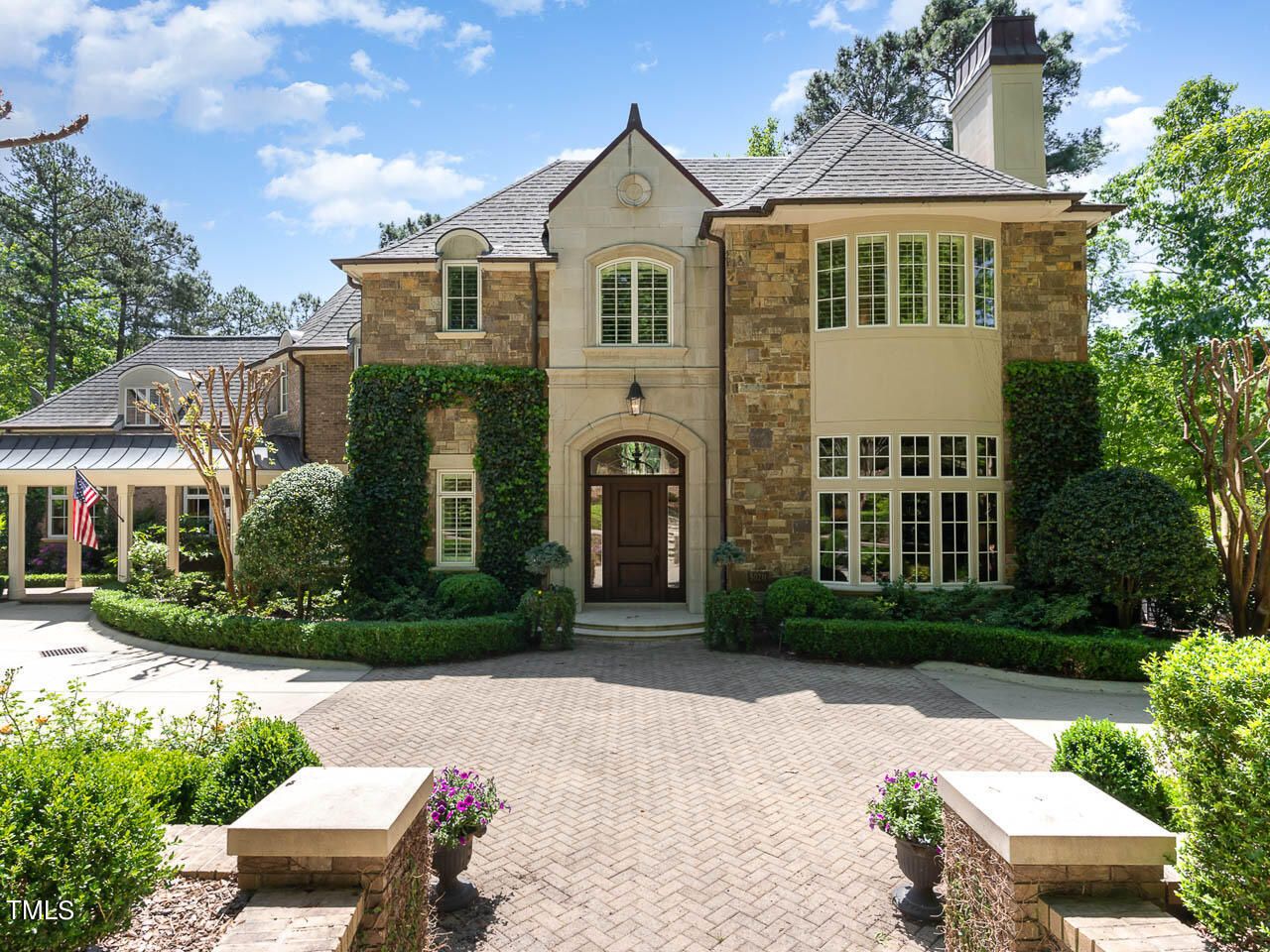 Front view of a large, elegant stone house with a dark wooden door, tall arched windows, green ivy on walls, manicured shrubs, and a brick driveway under a blue sky with clouds.