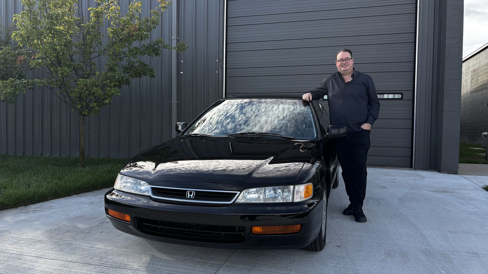 Man in a dark shirt and glasses stands with one hand on the roof of a black Honda sedan parked in front of a tall gray industrial garage door; a tree and concrete driveway frame the scene.
