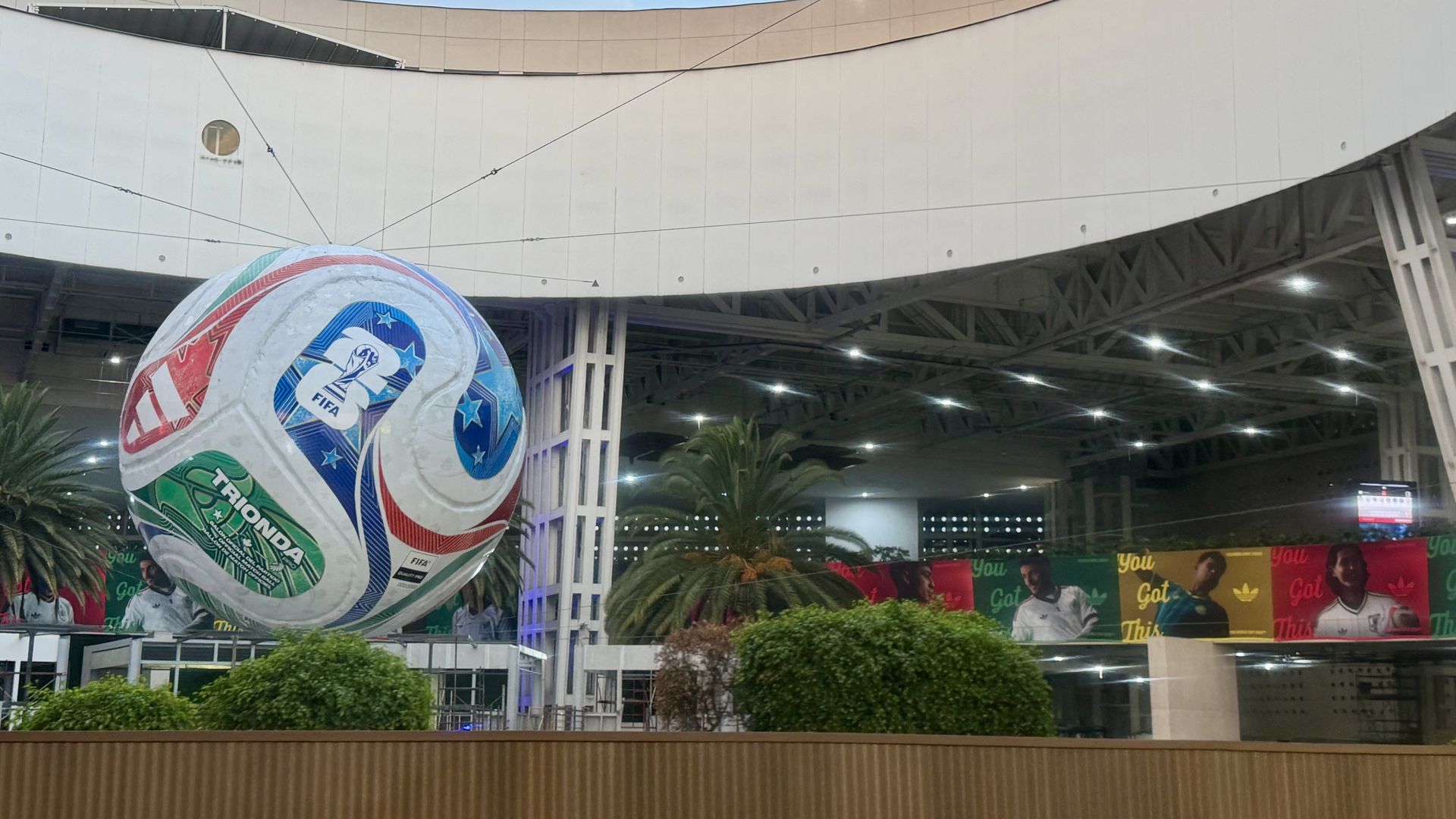 Indoor plaza with a giant FIFA soccer ball sculpture in the foreground, a curved white wall overhead, visible steel trusses, bright ceiling lights, and colorful banners with people along the lower wall.