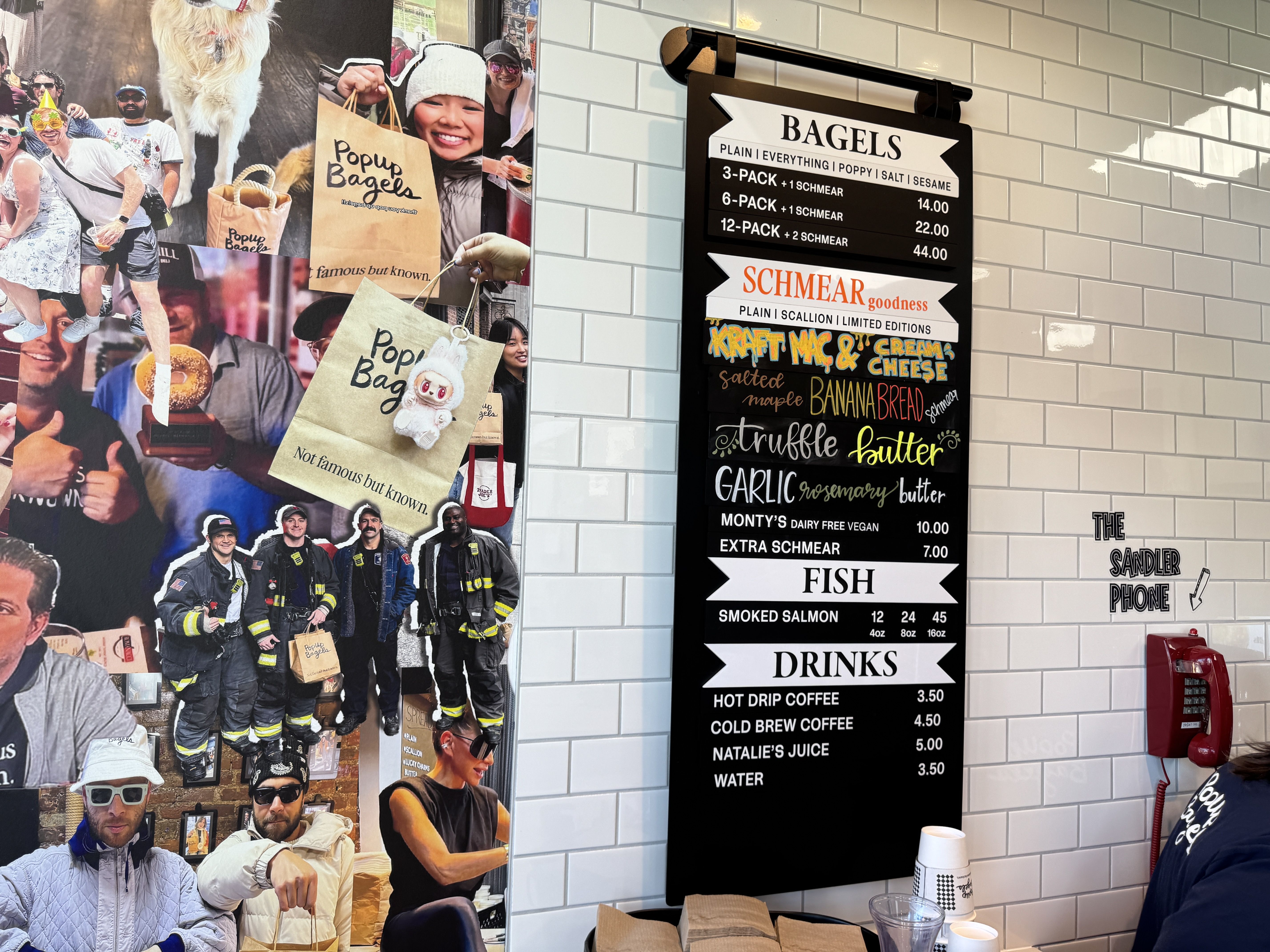 Bagel shop menu on white tiled wall listing bagels, schmear flavors, fish, and drinks; next to a red phone labeled "The Sandler Phone" and a collage of people holding Popup Bagels bags.
