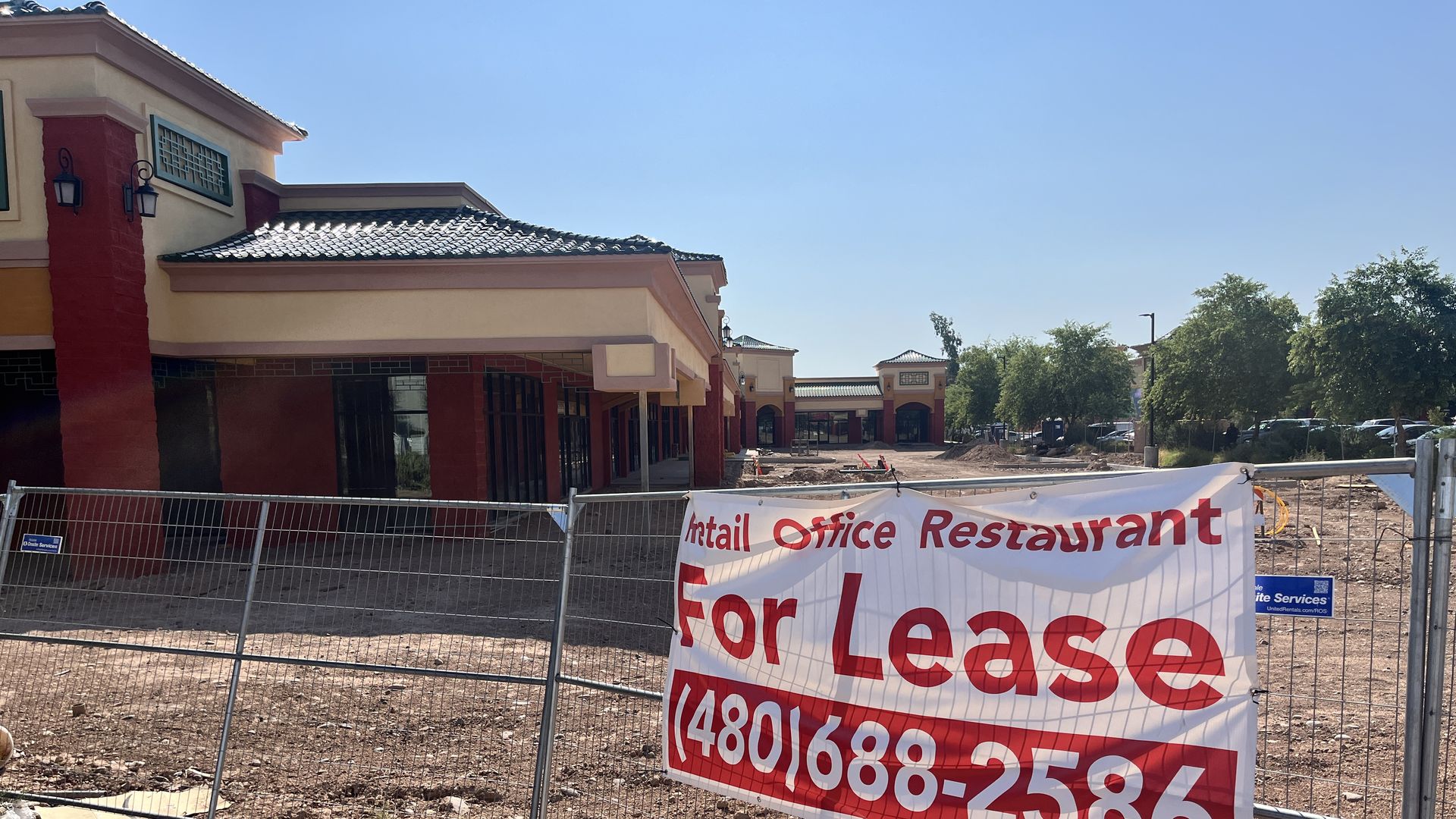 A sign that says, "For lease" with a strip mall that has a decorative roof int he background. 