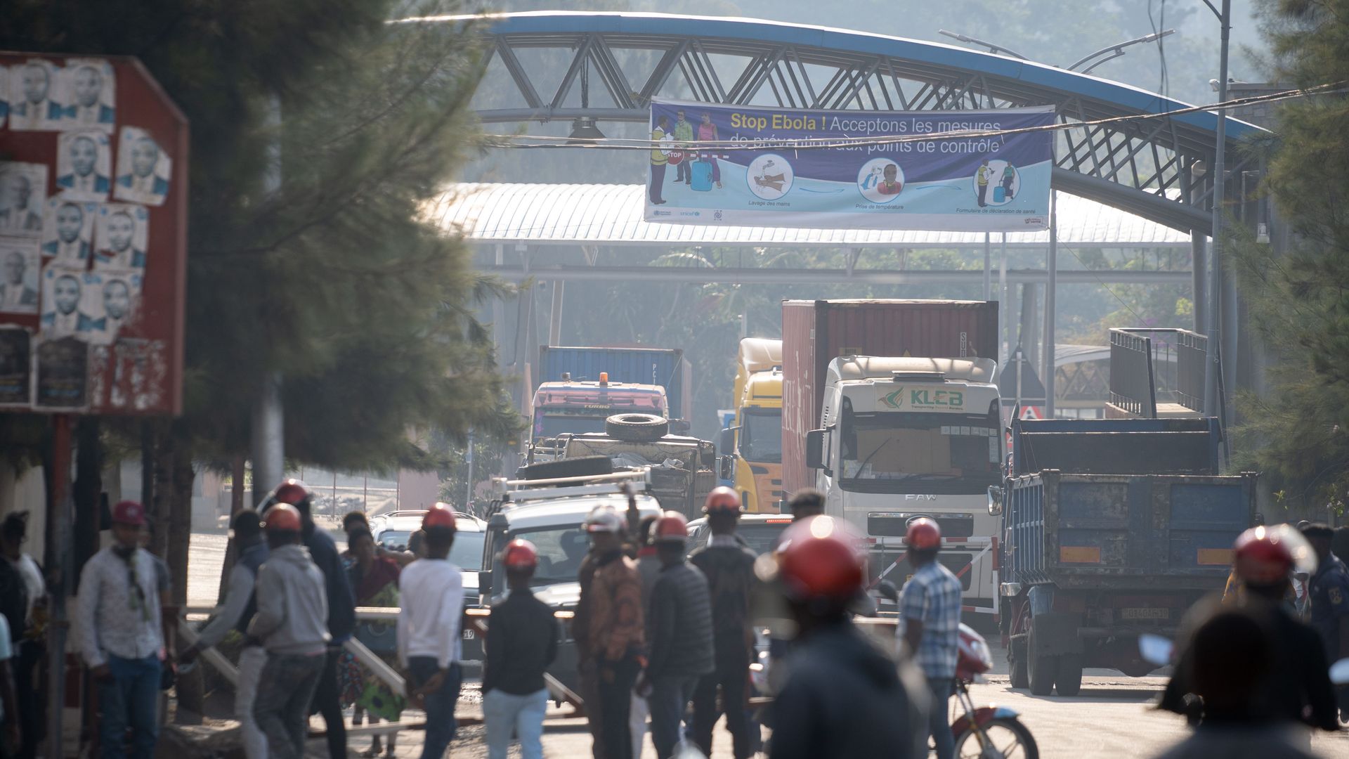 In this image, people on bikes and pedestrians walk on a dirt road with trucks in the background