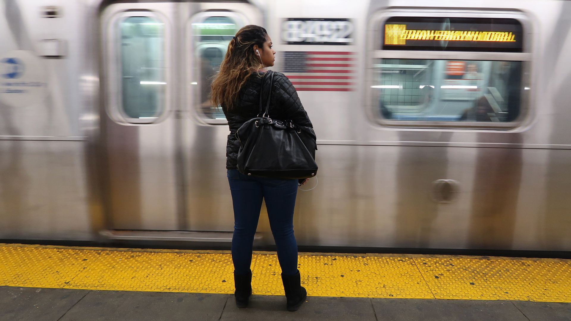A woman stands near the yellow strip on the concrete ground as a subway train goes past her. 