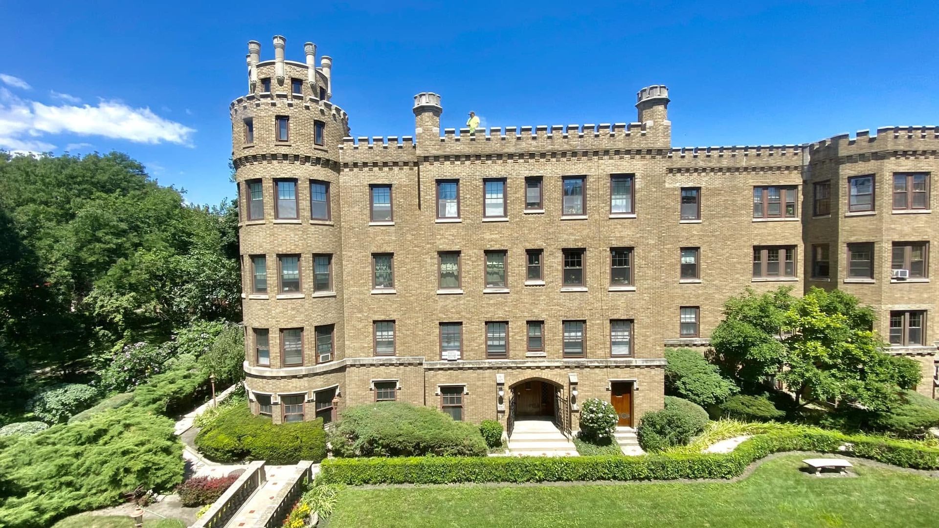 Castle-style brick building with crenellated towers under a bright blue sky. A person in a yellow shirt stands on the parapet, while green hedges and a manicured garden frame the foreground.