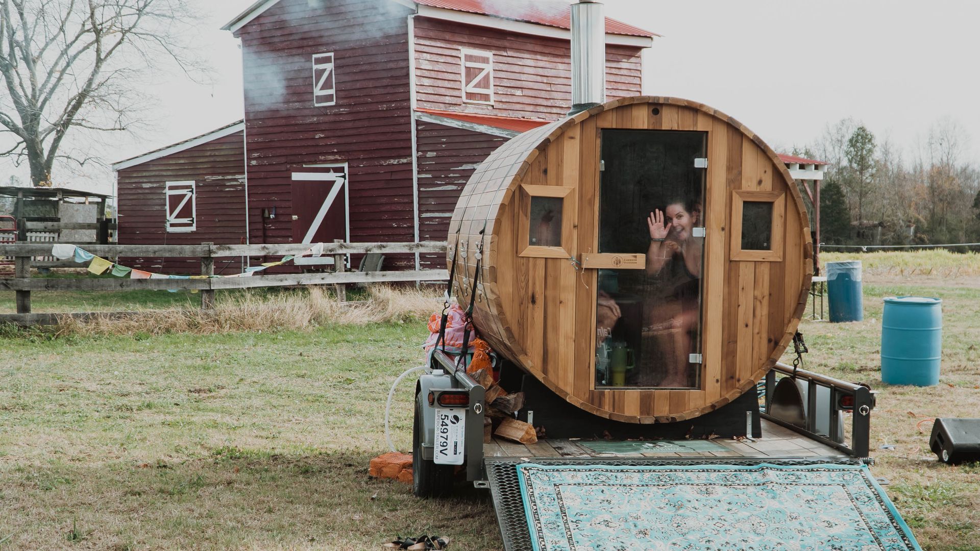 A pic of a farm and a red barn with a barrel-shaped sauna in front of it and a woman waving hello out of it