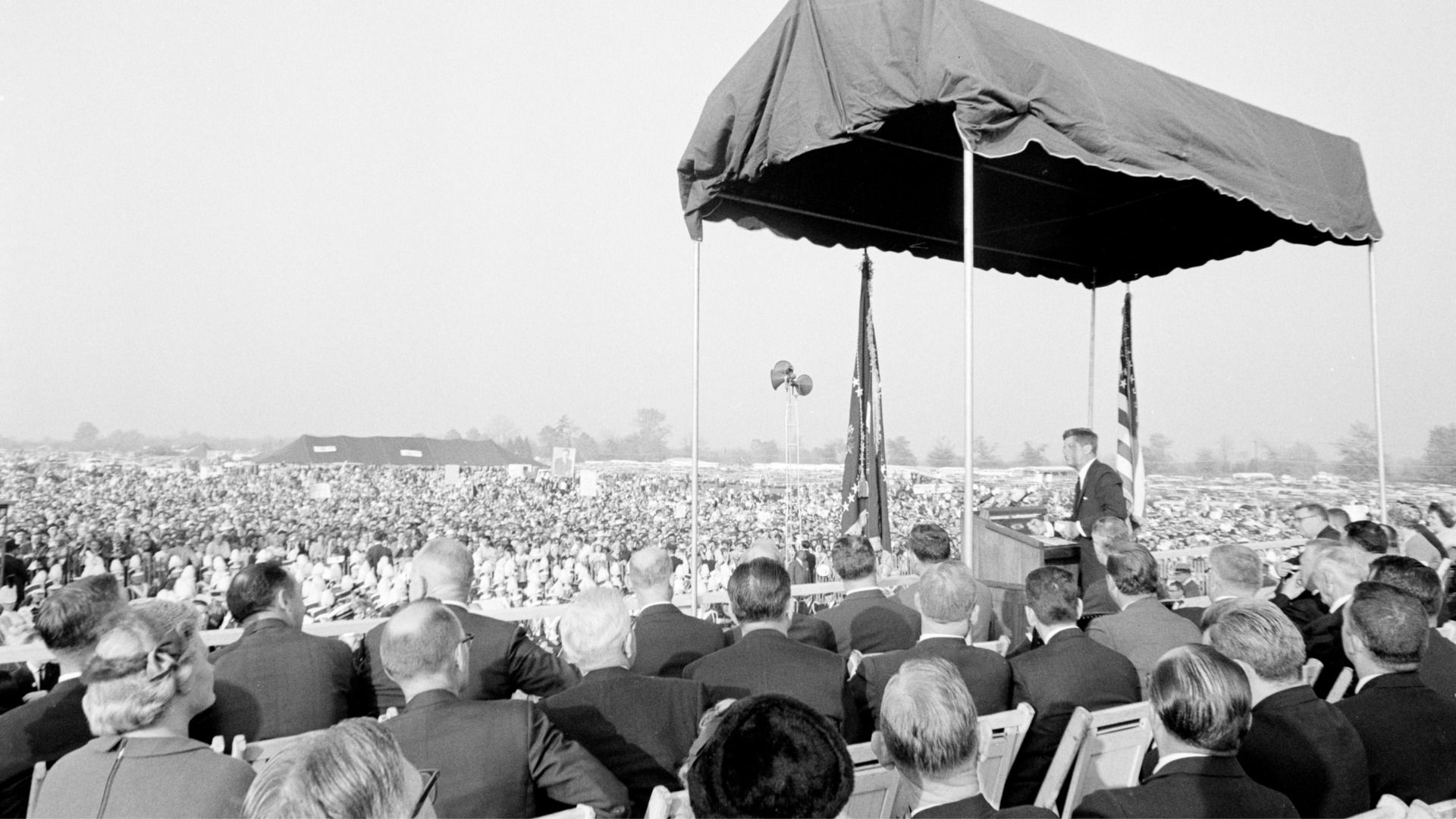 Black-and-white outdoor rally: a speaker at a podium under a canopy addresses a vast seated crowd, with the U.S. flag visible beside another flag.