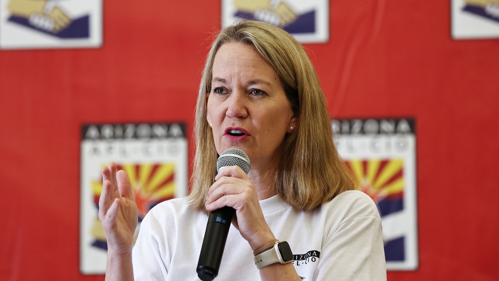 Blonde woman in white shirt speaking into a microphone with red background featuring Arizona AFL-CIO logos showing a handshake and starburst design.