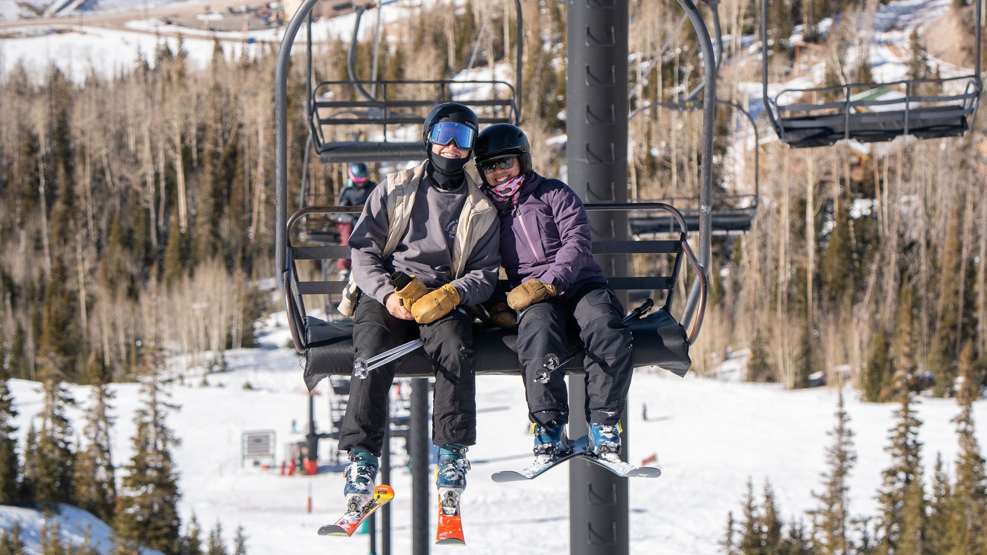 Two skiers wearing helmets and goggles sit close together on a ski lift above a snowy slope with trees in the background, enjoying a sunny winter day.