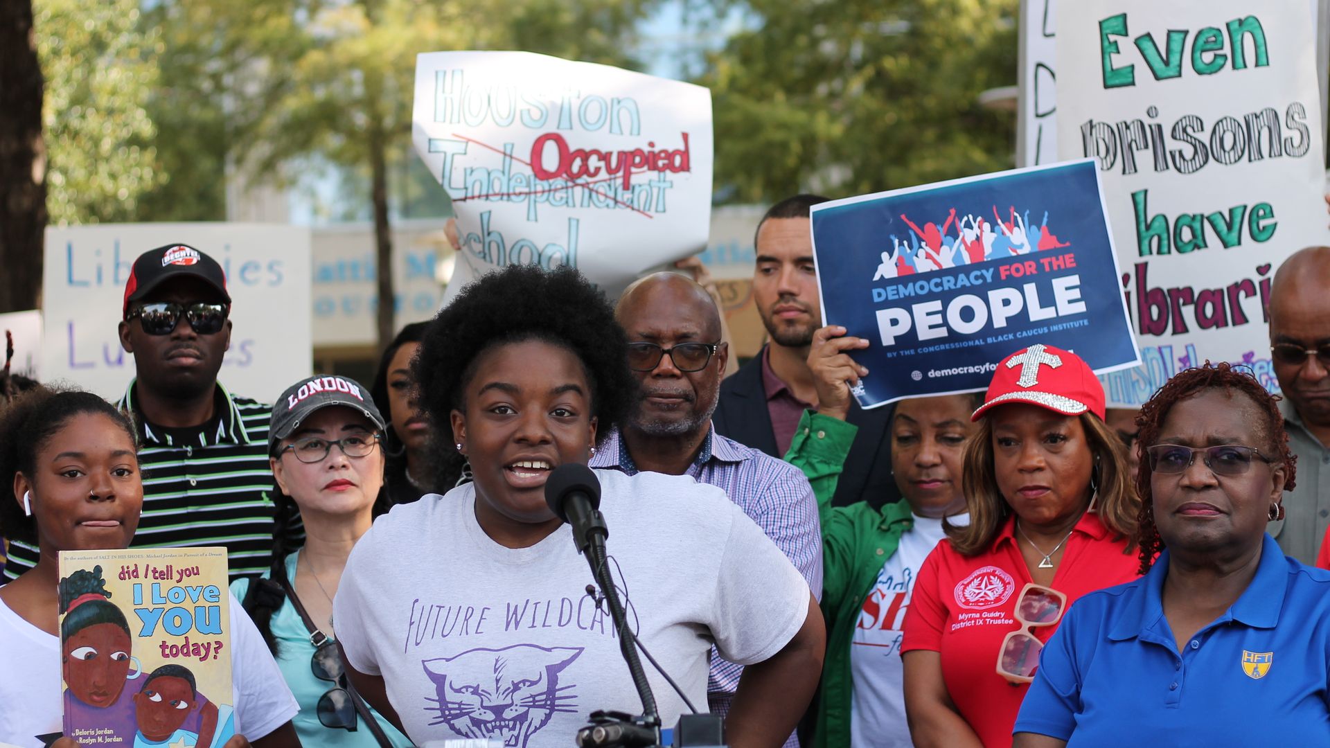 Photo of an 11th grader speaking at a rally, with people holding up handmade signs. 