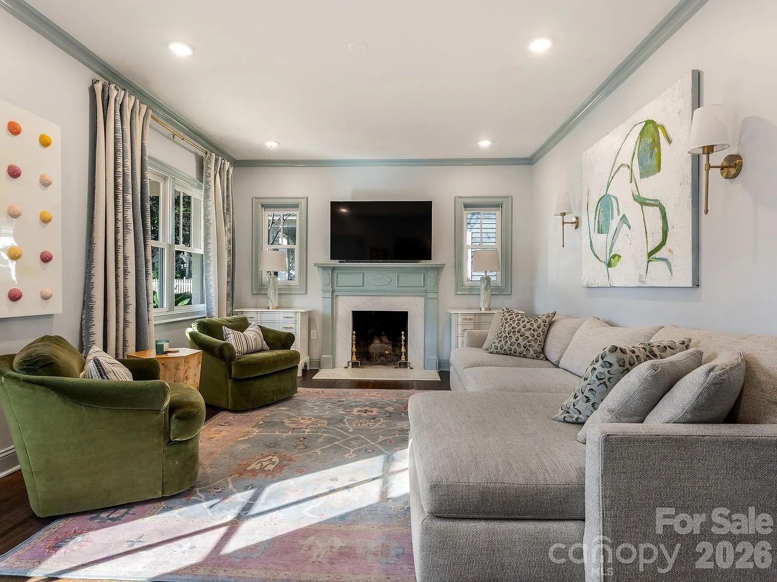 Living room with gray walls, green velvet armchairs, a gray sectional sofa, a fireplace with a mounted TV, two windows with curtain panels, abstract green-and-blue painting, patterned rug.