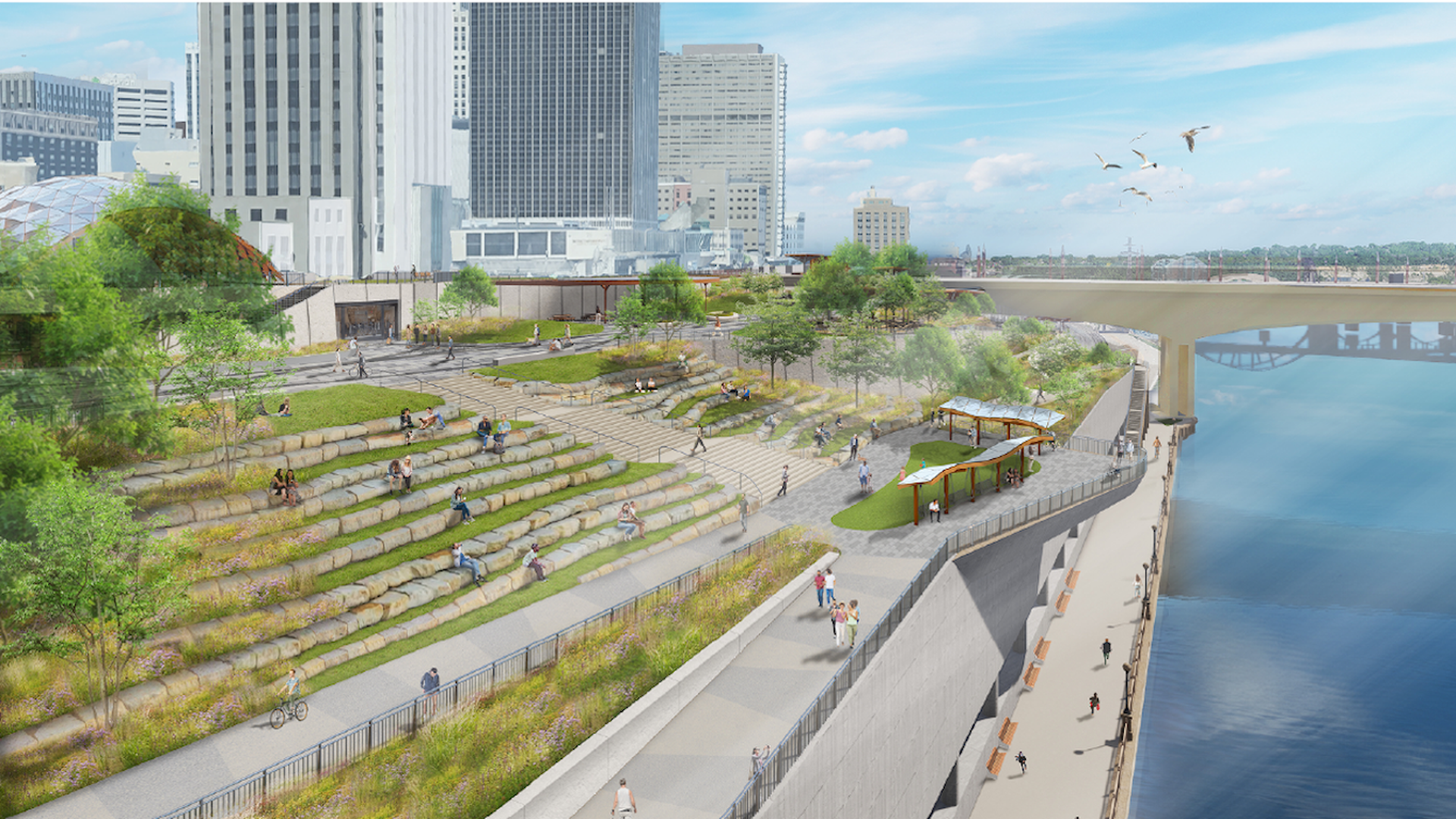 Urban riverside park with terraced grassy steps, trees, and winding paths along a blue river. Modern high-rises in the background, a bridge, and people strolling, biking, and sitting by the water.