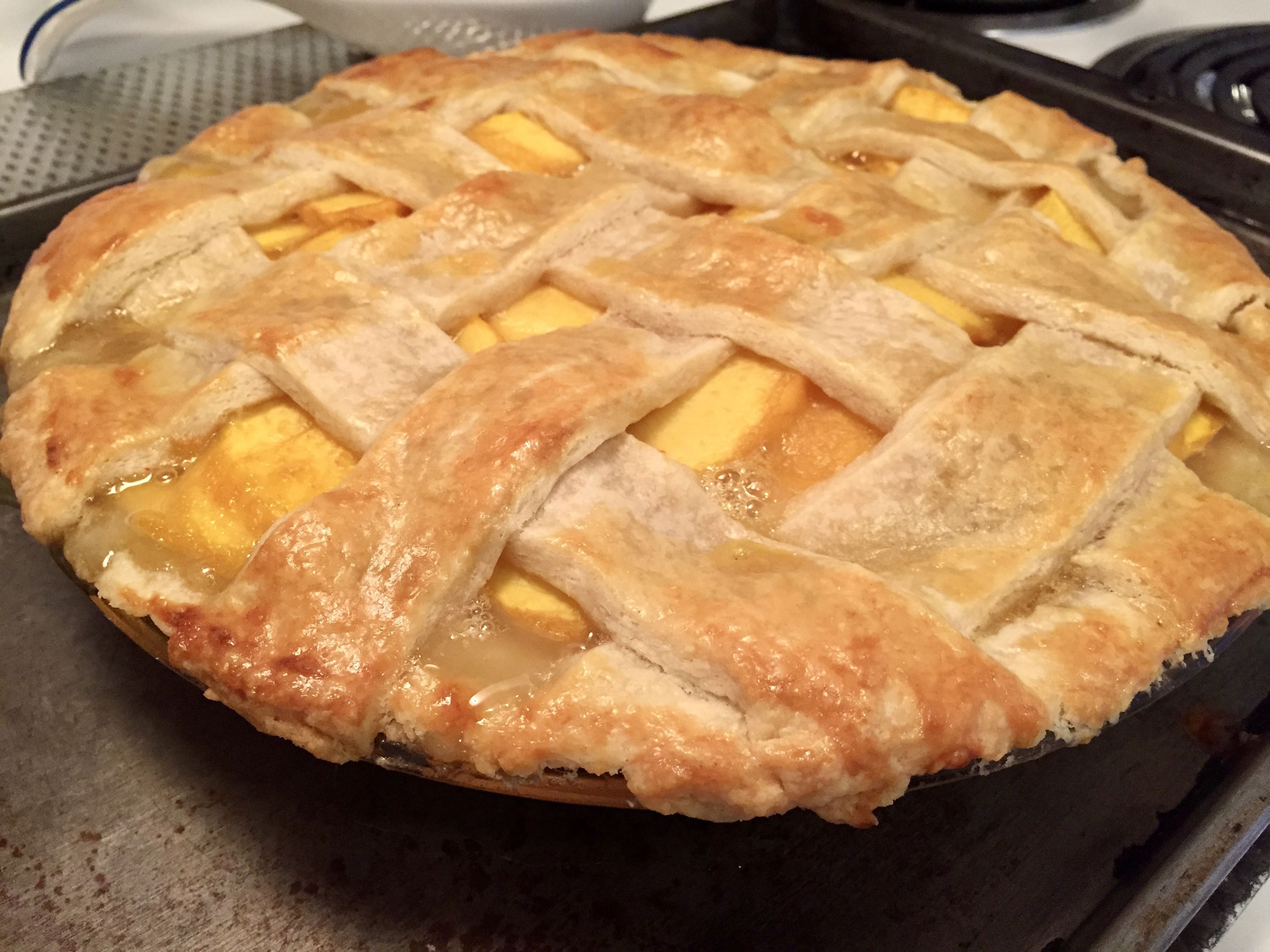 Golden-brown lattice crust peach pie with bubbling filling in a glass pie dish on a baking sheet next to a stove.