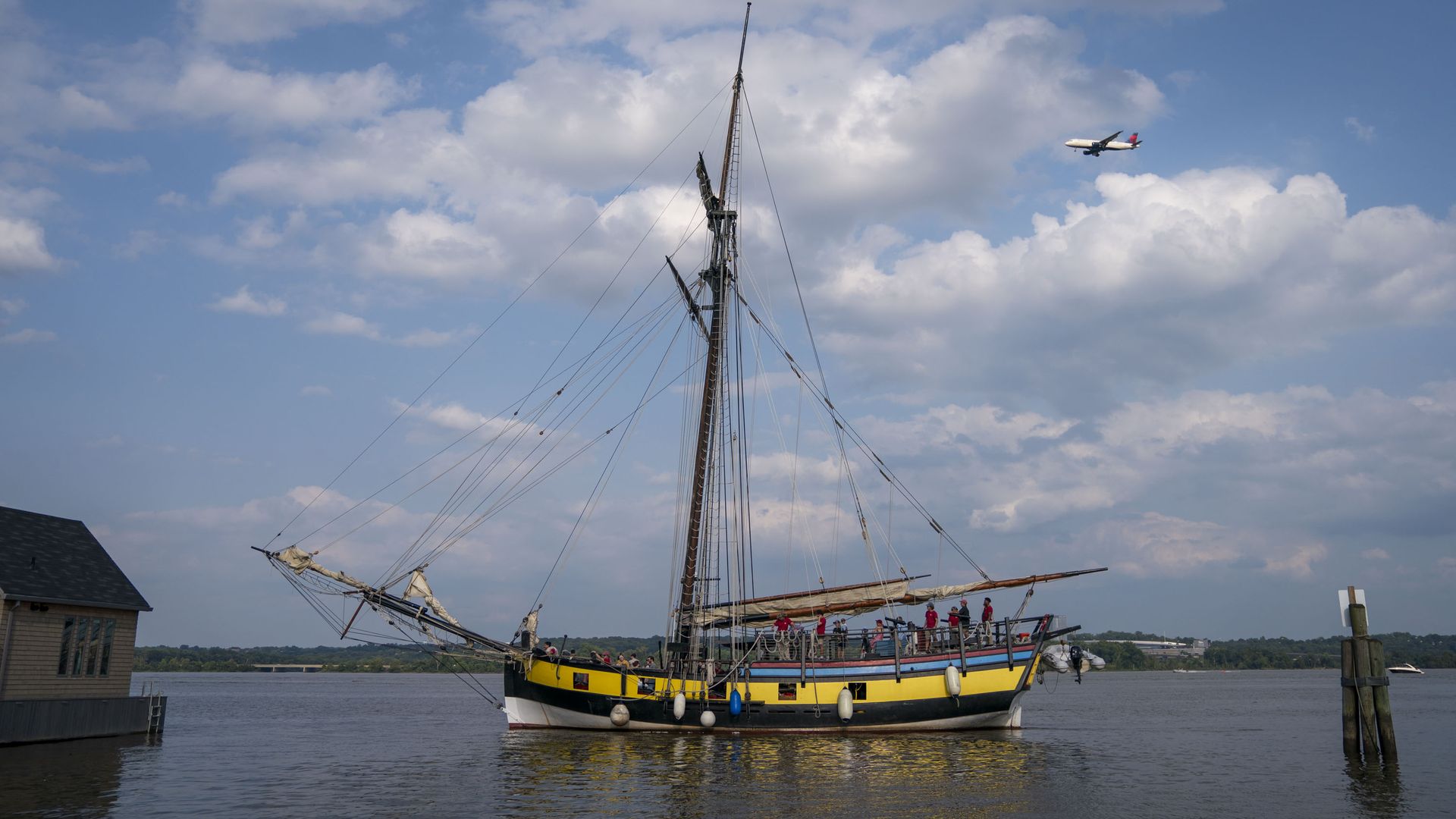 A colorful tall-ship with a yellow and black hull sits at a dock, its masts and rigging rising above calm water; people stand on deck. A small airplane flies in a blue sky with white clouds.