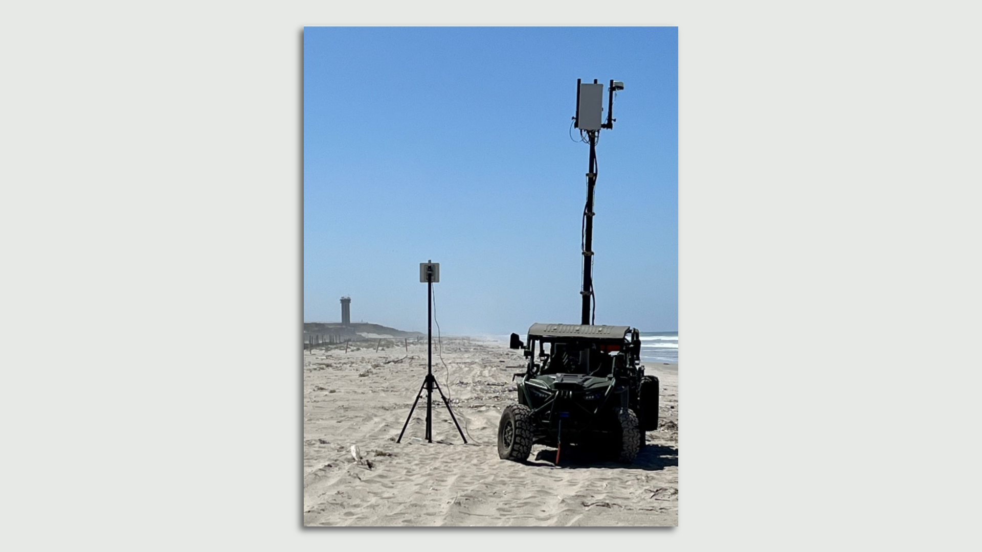 An all-terrain vehicle is parked on the beach. A small cell tower extends from the back of it. Blue skies are seen in the background.