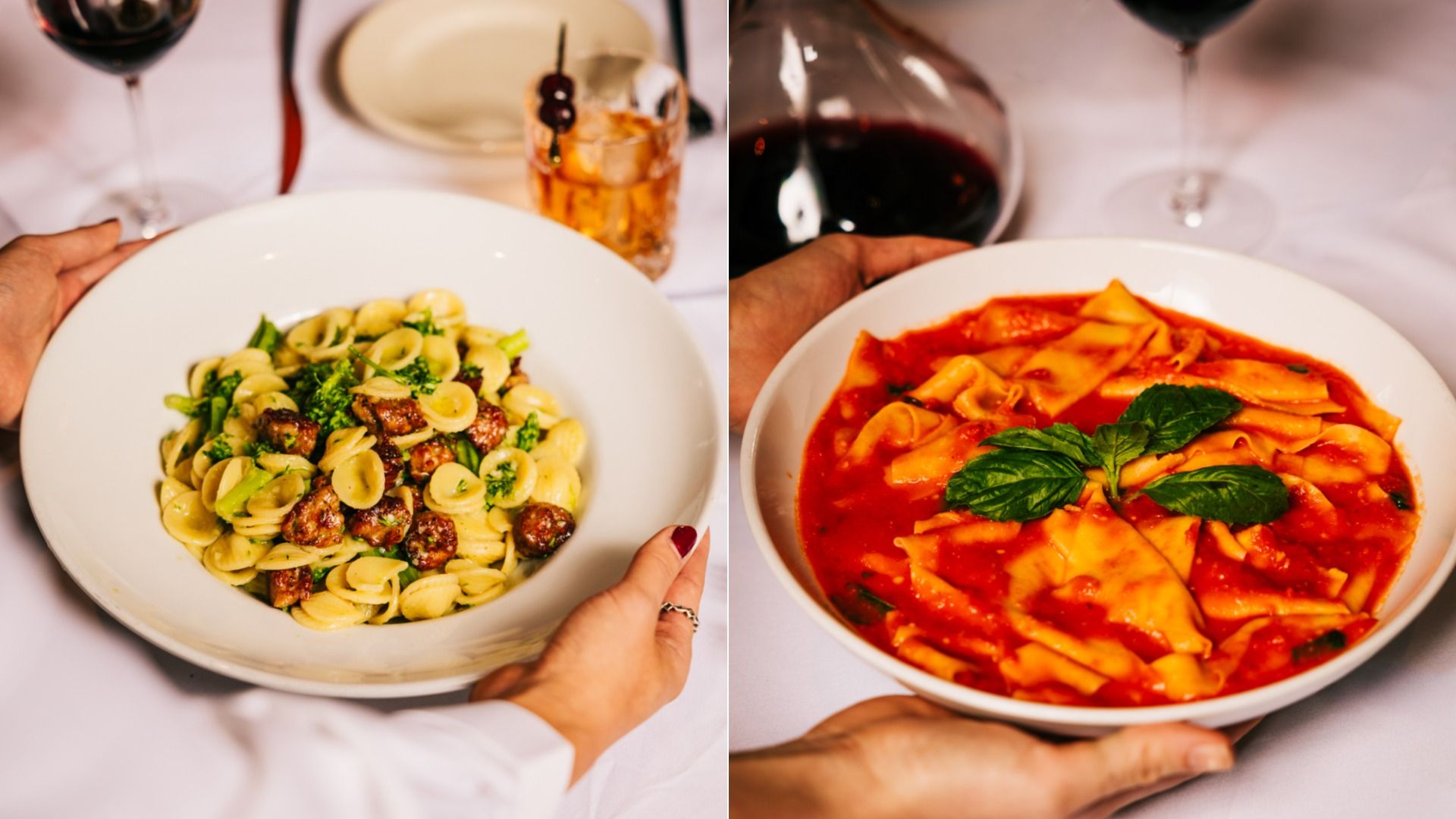 Two hands hold white bowls of pasta: left bowl has orecchiette with greens and sausage, right bowl has wide pasta in red tomato sauce topped with basil. Background includes drinks and white tablecloth.