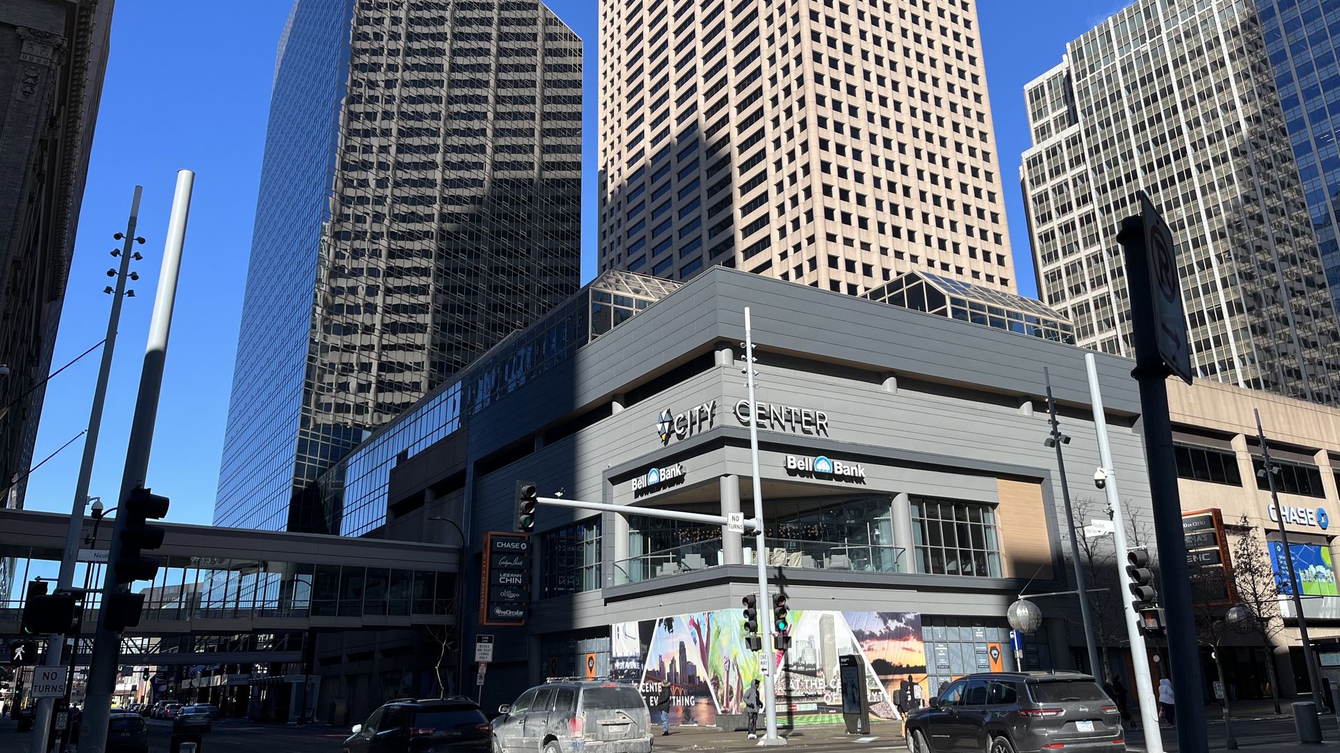 Urban street scene with tall skyscrapers under a blue sky: a triangular dark-glass tower beside a beige grid building, a glass office block to the right, and a "City Center" sign with traffic.