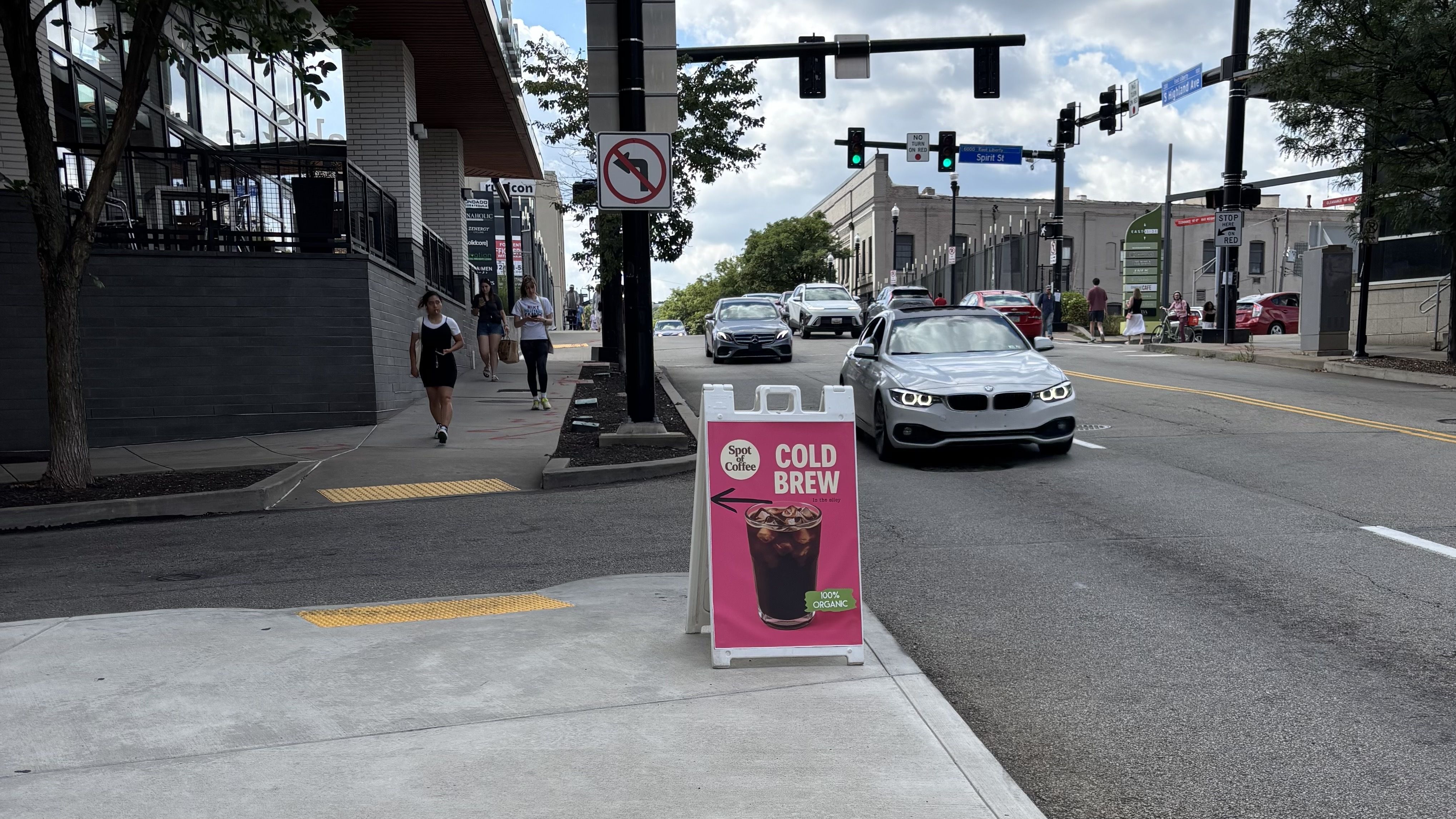 Street scene with pedestrians walking on sidewalk, several cars on road, traffic lights, and a pink sign advertising 100% organic cold brew coffee pointing left to Spot of Coffee.