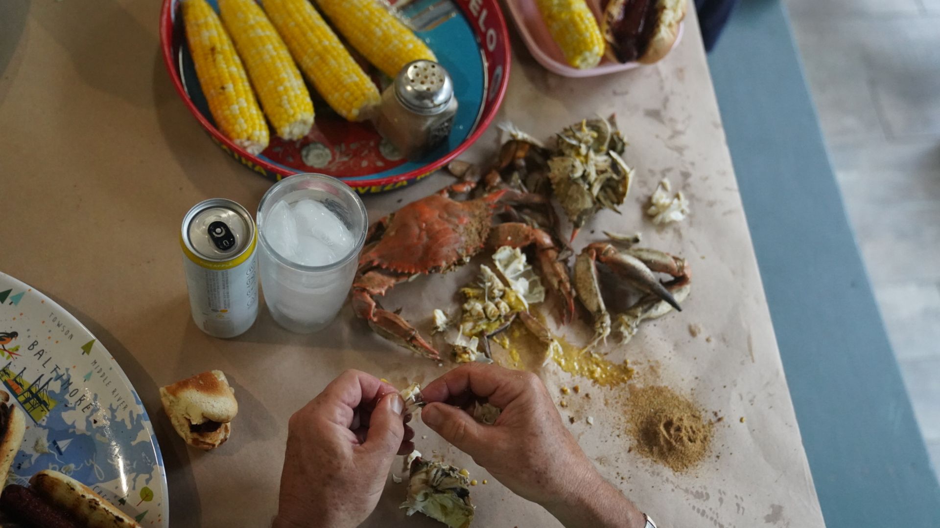 Hands cracking crab legs on a paper-covered table with cooked crabs, corn on the cob in a Modelo tray, iced drink can, glass of ice, hot dogs on plates, and seasoning pile.