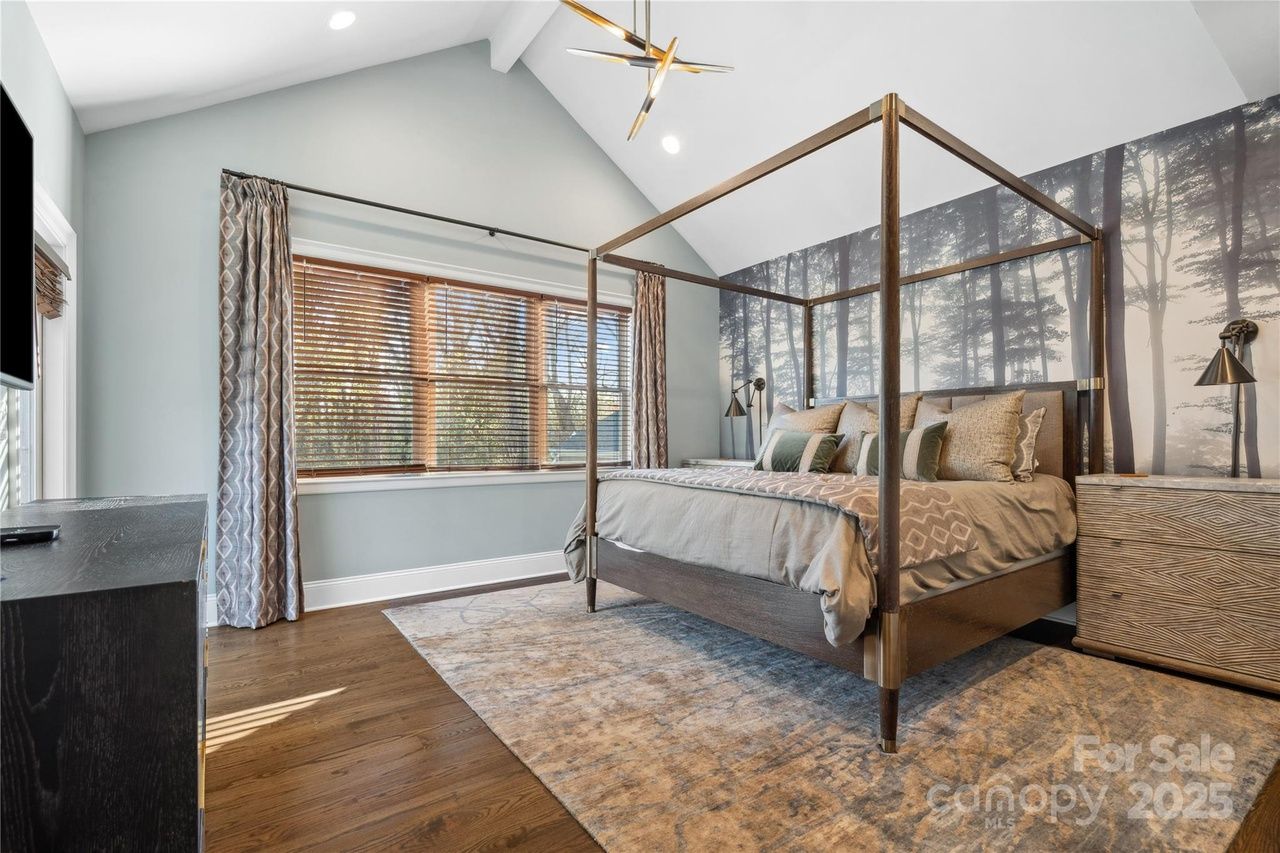 Modern bedroom with vaulted ceiling, wood and gold four-poster bed, gray and beige bedding, forest mural wall, patterned curtains, wooden blinds, wooden floor, and area rug.